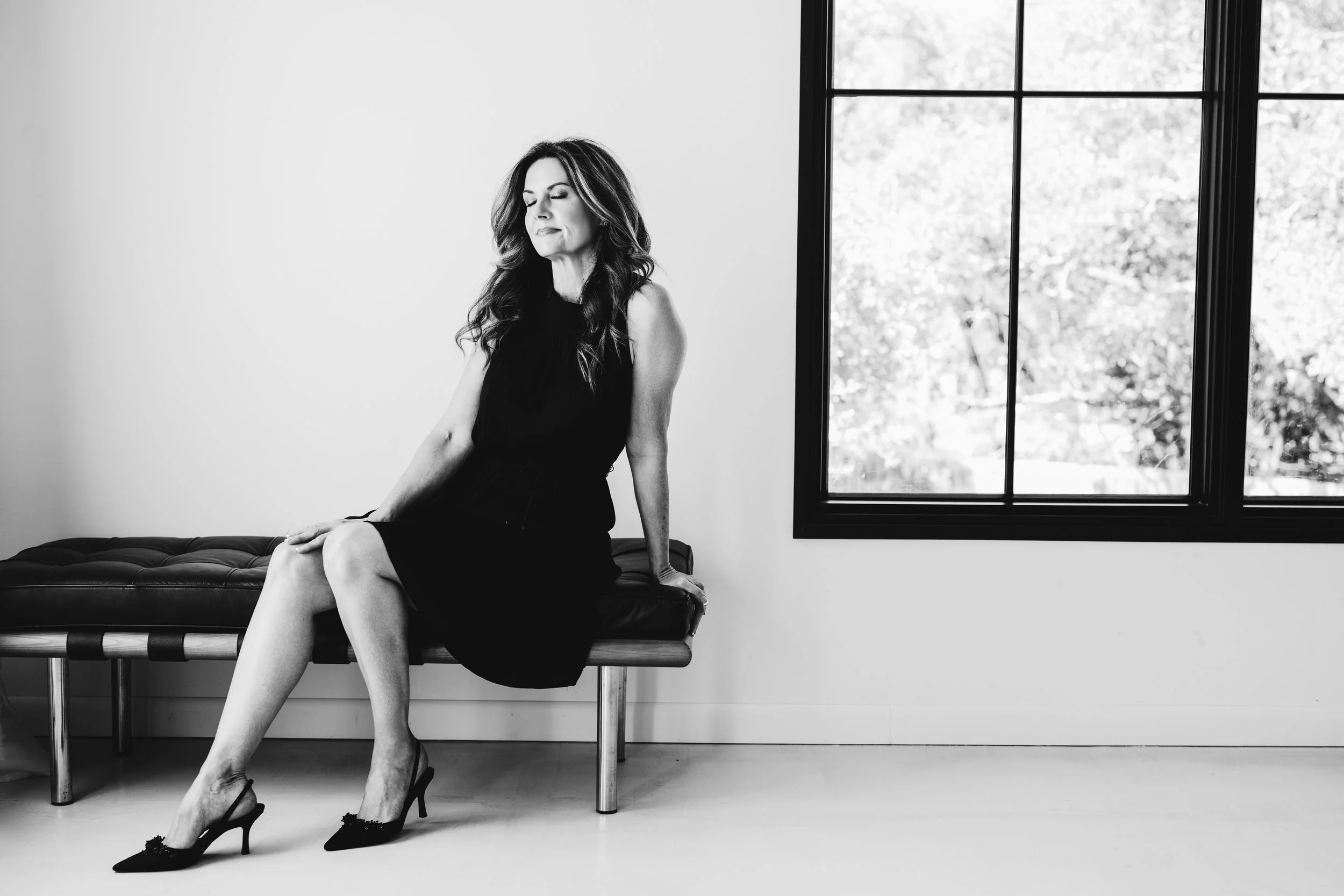 Elegant black and white portrait of a woman sitting on a bench beside a large window