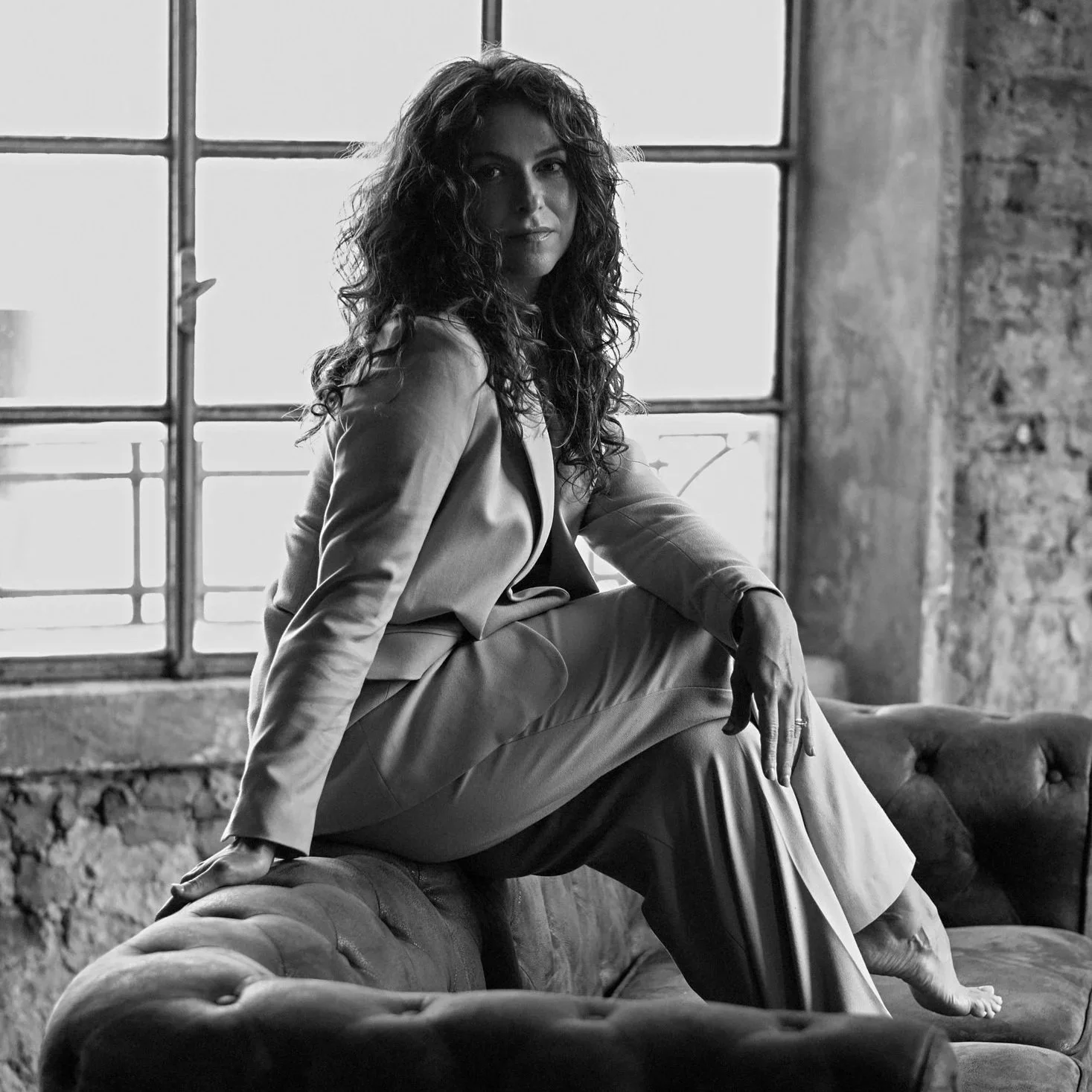 A woman with curly hair sitting on the armrest of a vintage leather sofa in front of a large window, in a black and white photo.