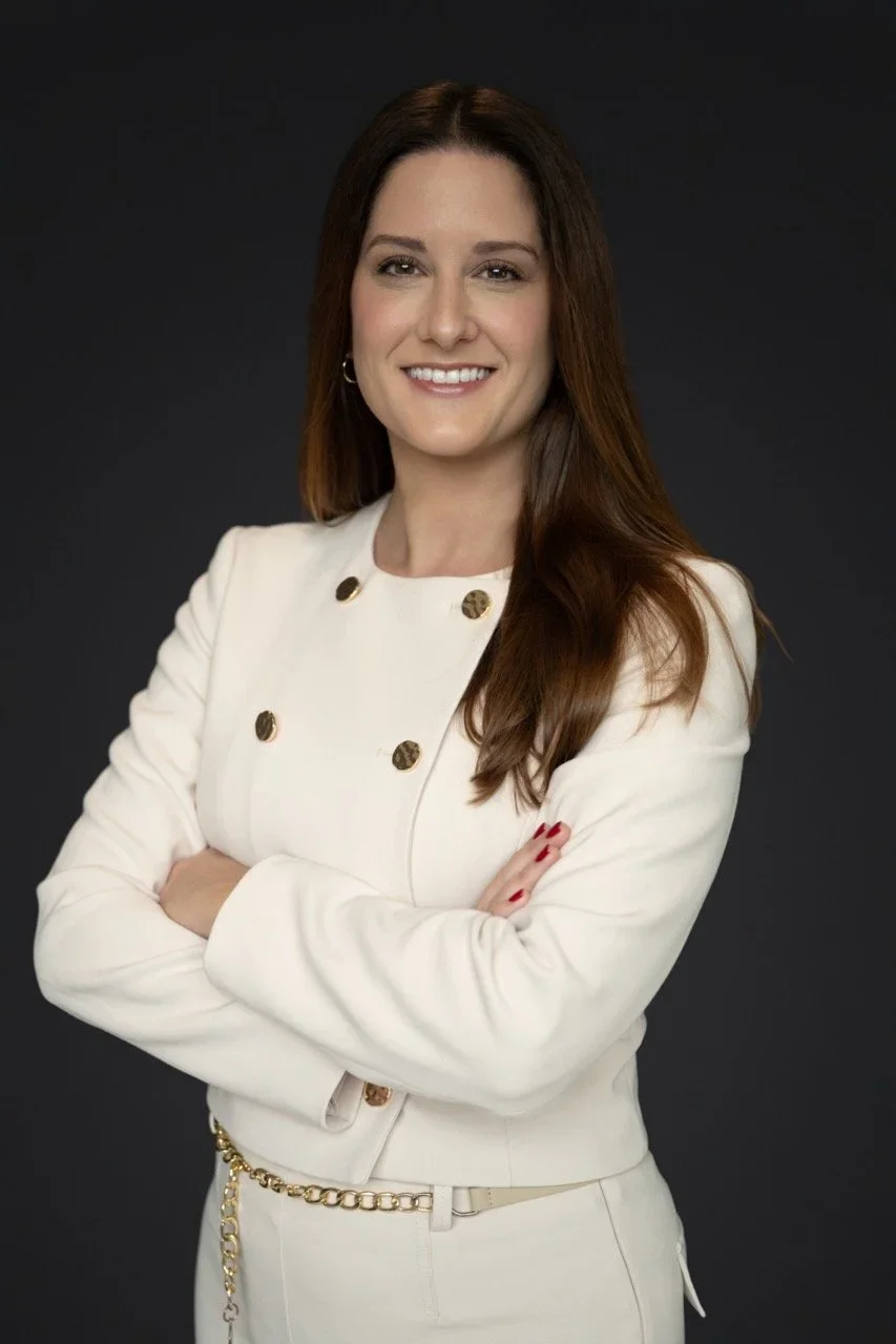 A woman with long brunette hair smiling, wearing a white blazer with gold buttons and a matching skirt, posing with crossed arms against a dark background.