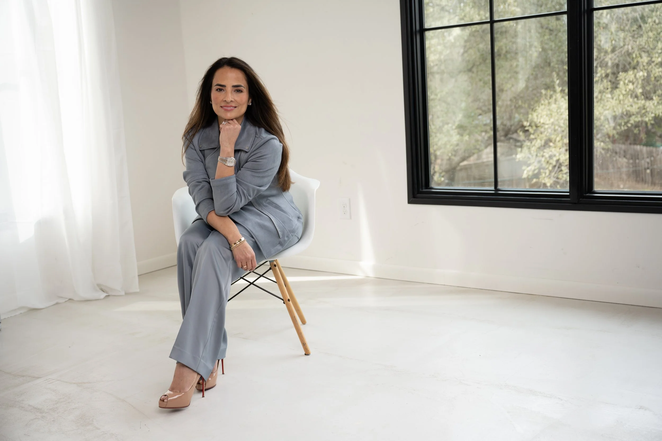 Professional branding portrait of a confident woman in a gray suit seated on a chair in a bright modern studio with natural window light, relaxed yet authoritative pose.