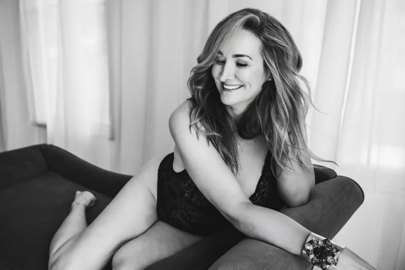 Black and white boudoir portrait of a smiling woman in black lace lingerie reclining on a studio chaise