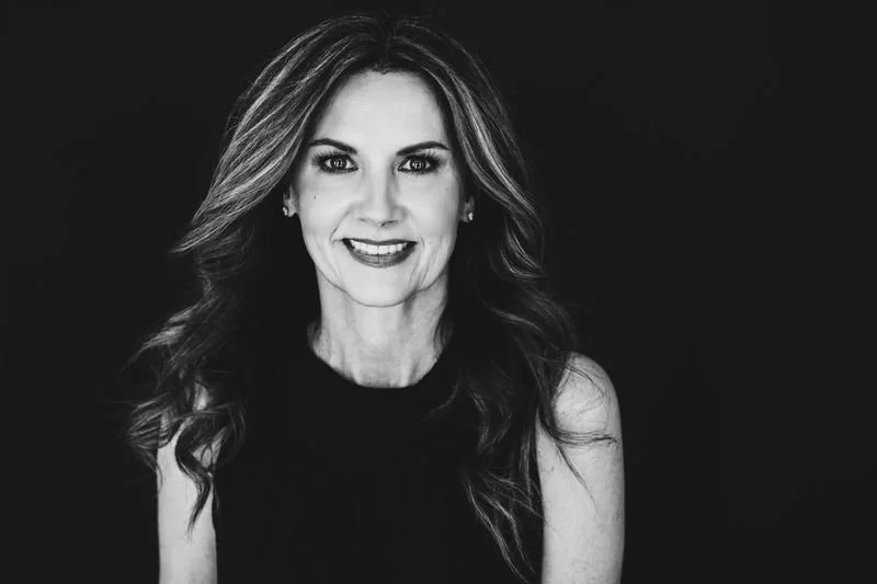 Black and white portrait of a smiling woman with long wavy hair, wearing a sleeveless top, against a dark background.