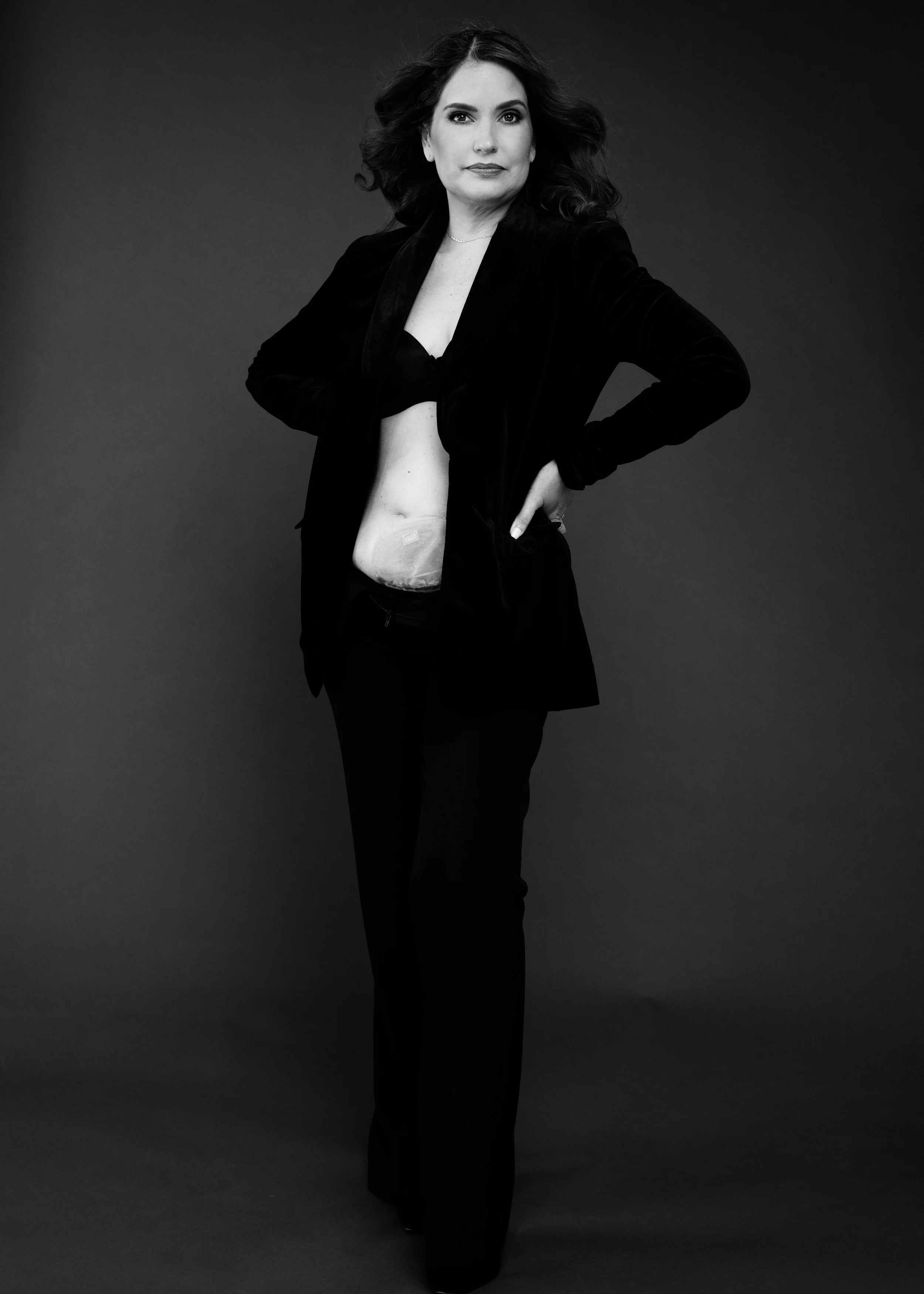 A woman with long wavy hair posing confidently in a black outfit with a bra and partial stomach exposed, against a dark background.