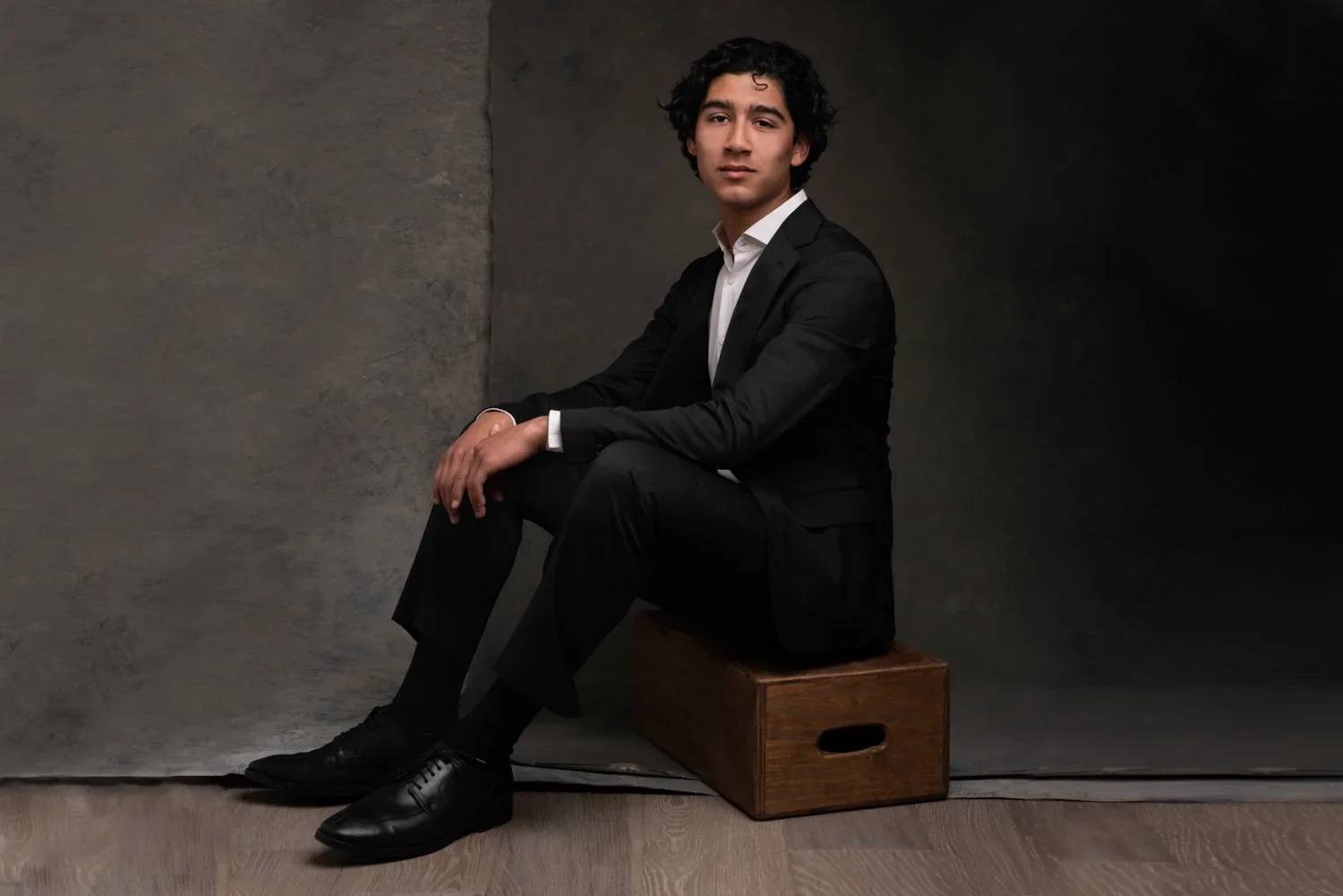 A young man in a black suit sitting on a wooden box, against a dark background.