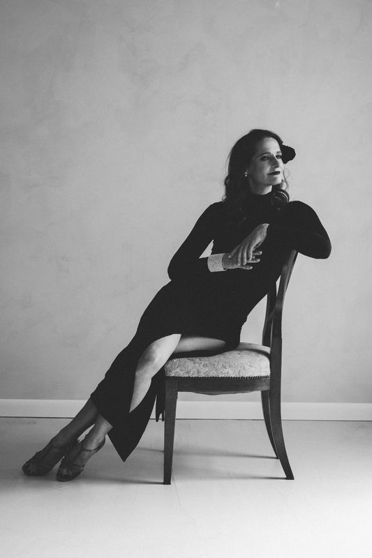 A woman in an elegant black dress sitting sideways on a wooden chair with one arm resting on the backrest and the other on her lap, looking to the right.