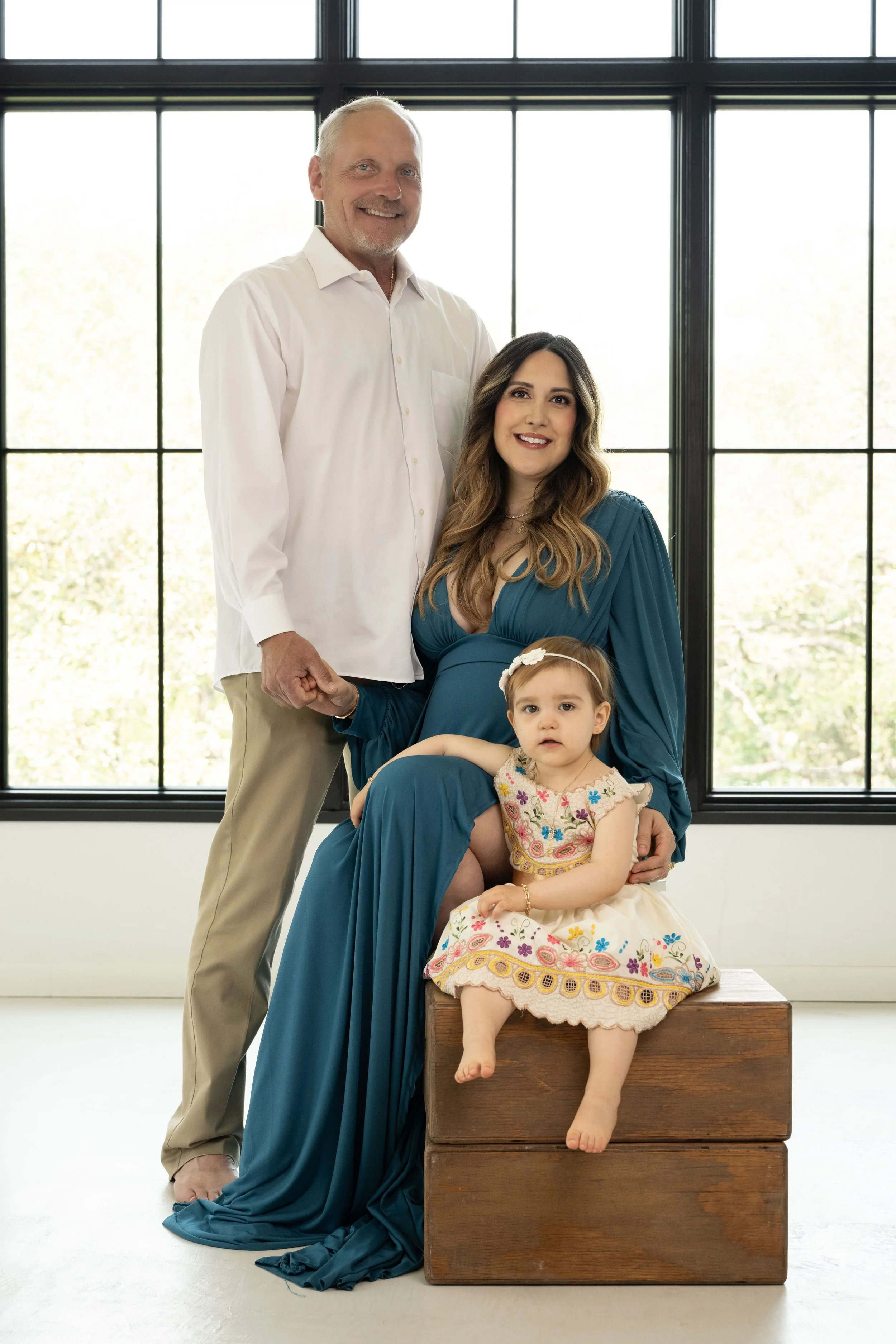 Family portrait of parents and young daughter seated by large studio window in natural light