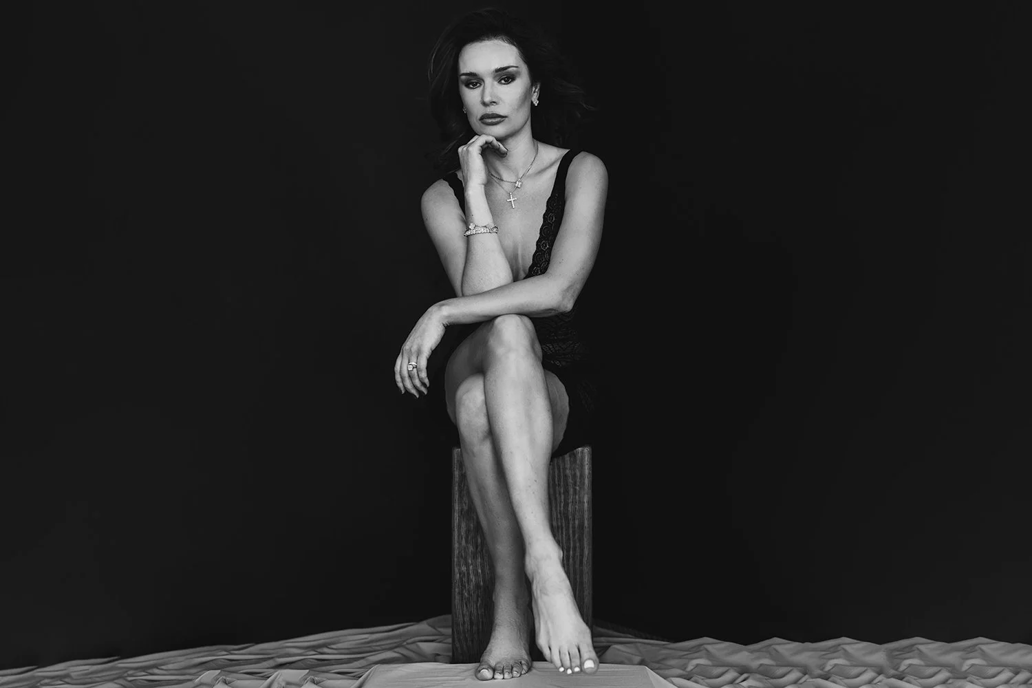 A black-and-white portrait of a woman seated with ease and intention. Her posture is relaxed yet commanding, legs crossed, shoulders open, gaze steady. There is no performance here only presence. The simplicity of the setting draws attention to her c