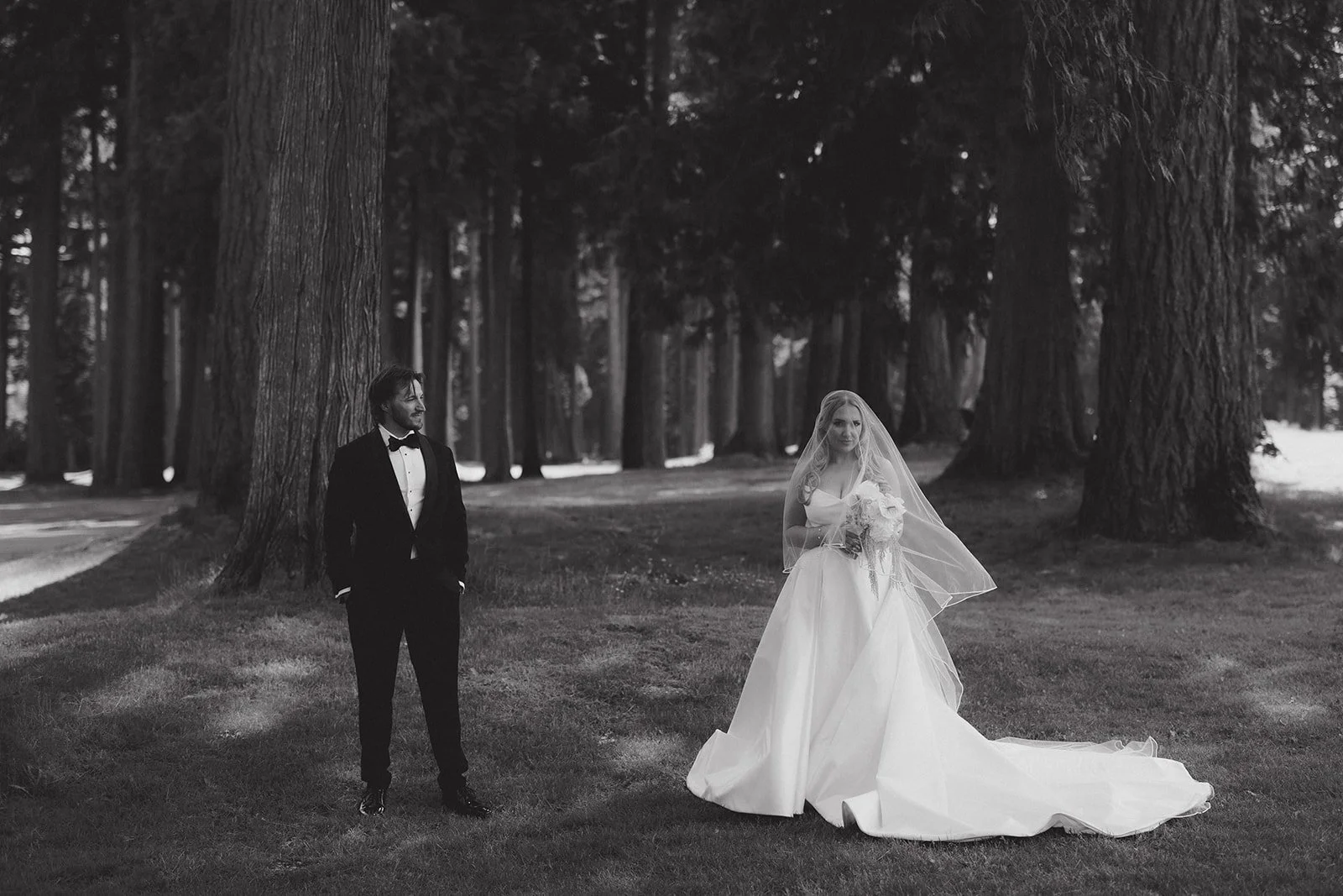 A bride in a wedding gown holding a bouquet and a groom in a tuxedo standing apart in a forest with large trees.