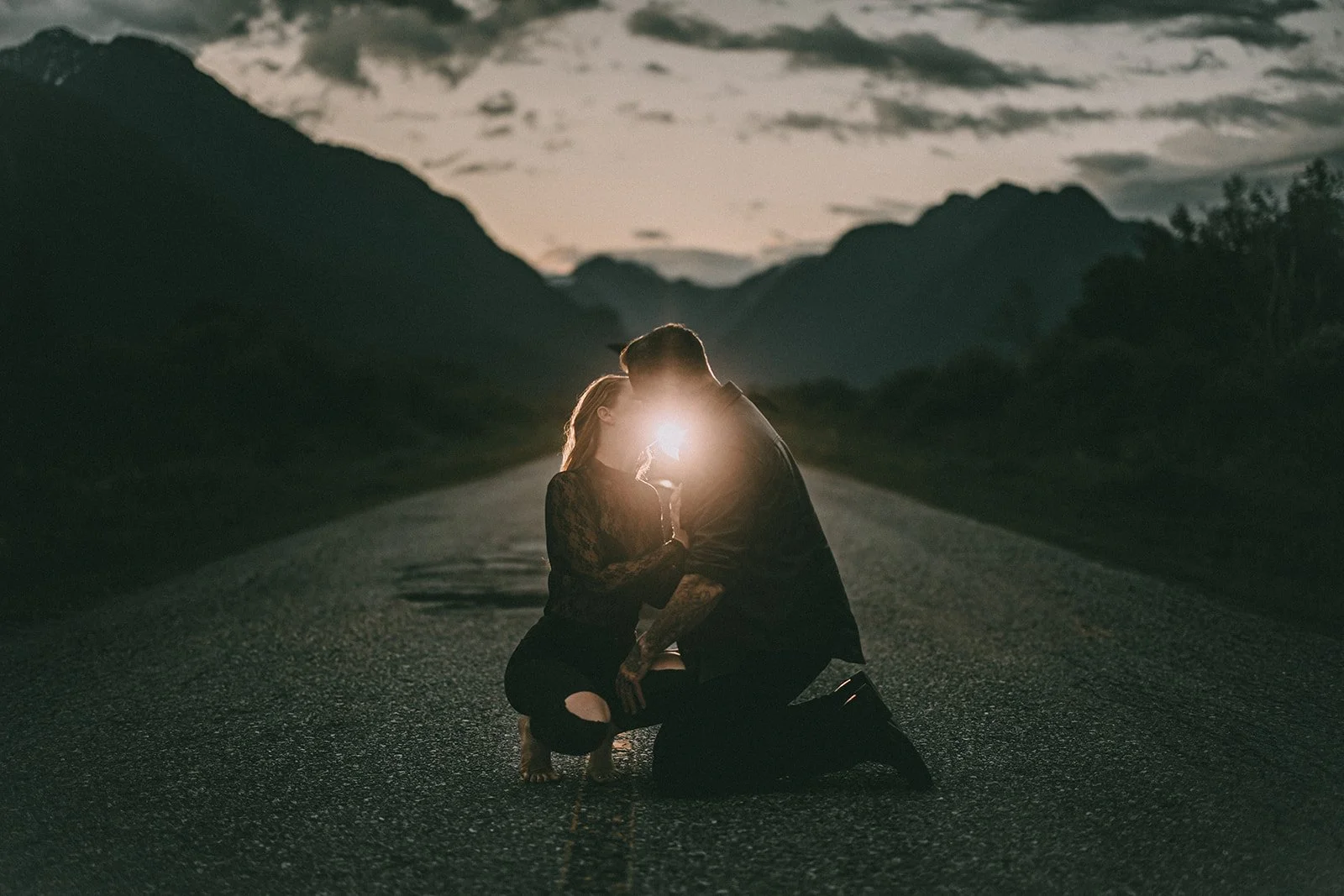 A couple is kneeling on an empty road at sunset or dusk, with mountains and cloudy sky in the background. They are close together, kissing, with the woman's face illuminated by the sunlight behind them.