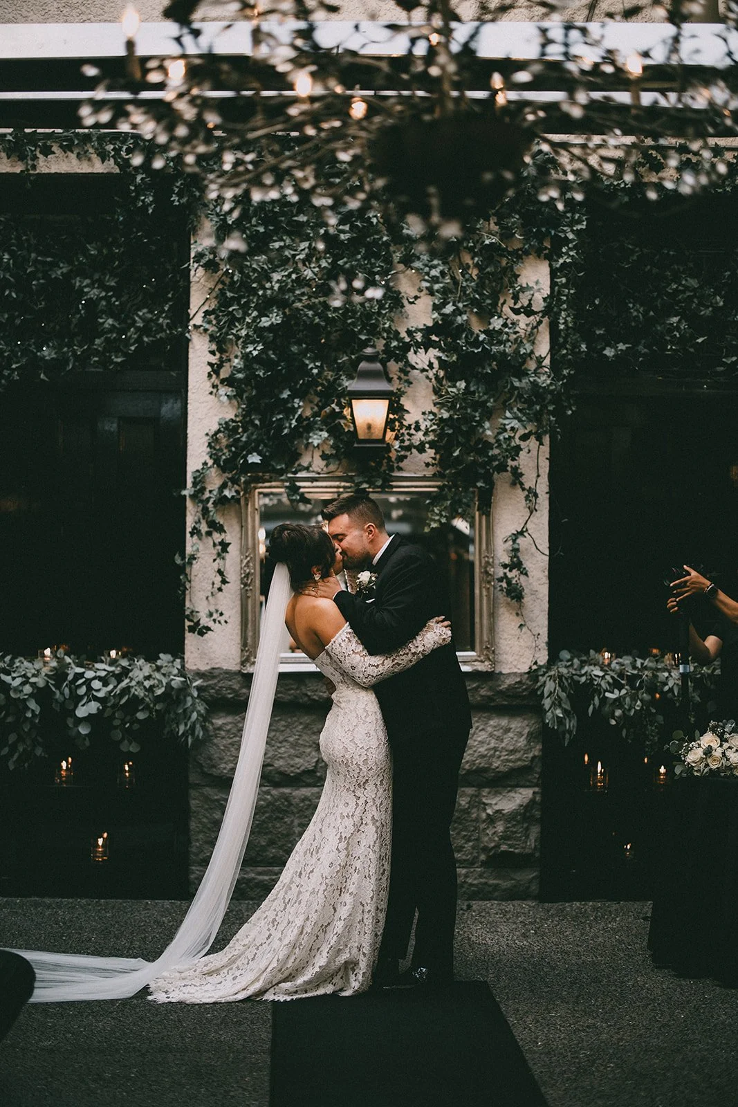 Bride and groom kissing at wedding, surrounded by ivy and candles, with chandelier above.