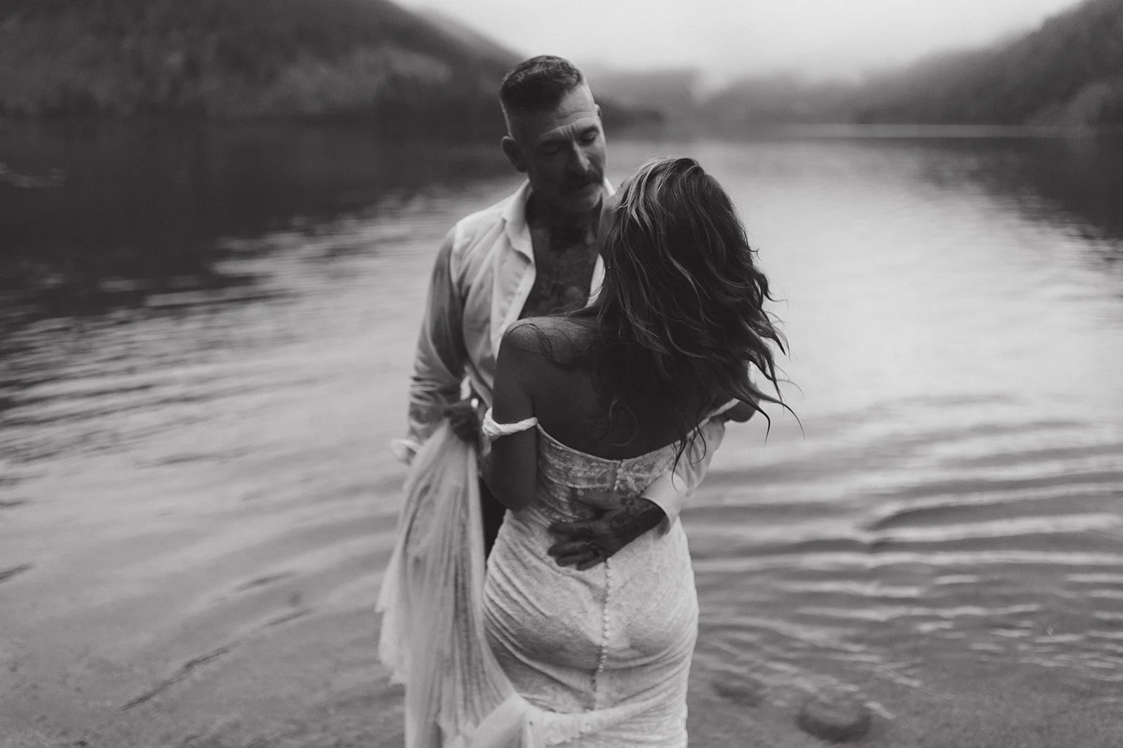 A black and white photo of a man and woman standing in a body of water, with rocky hills in the background, sharing an intimate moment.
