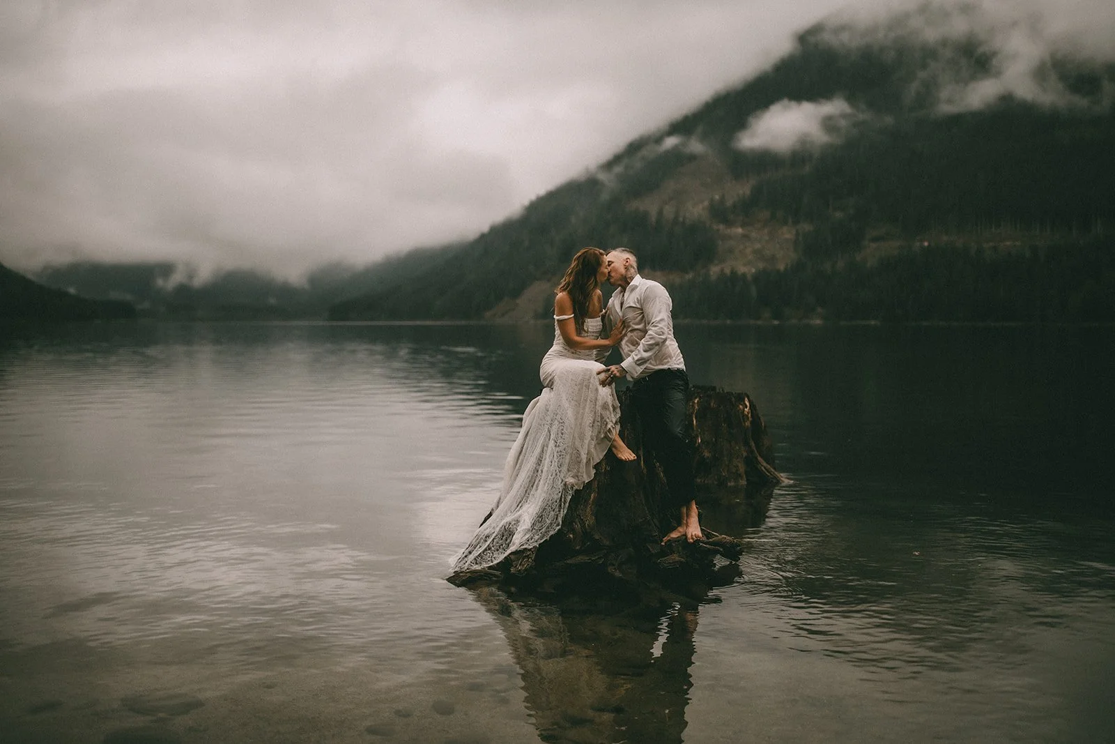 A couple dressed in wedding attire sitting on a tree stump in a lake, surrounded by mountains and cloudy sky, sharing a kiss.