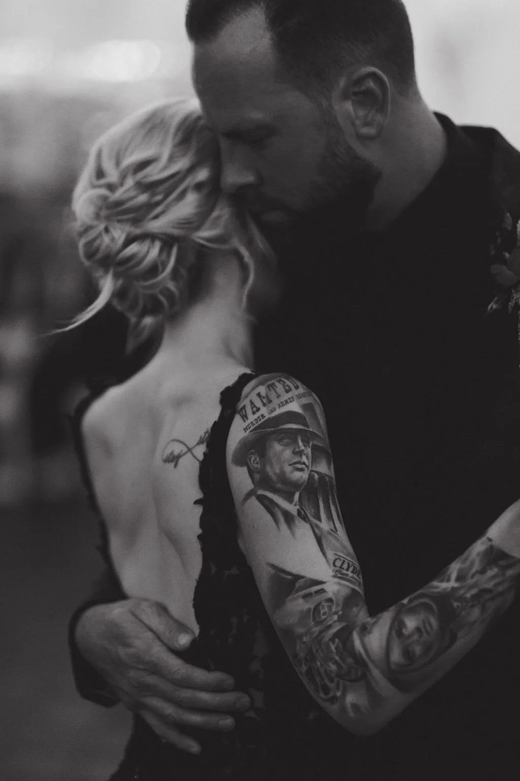 Black and white photo of a couple embracing, showing a woman's back with detailed tattoos on her arm.