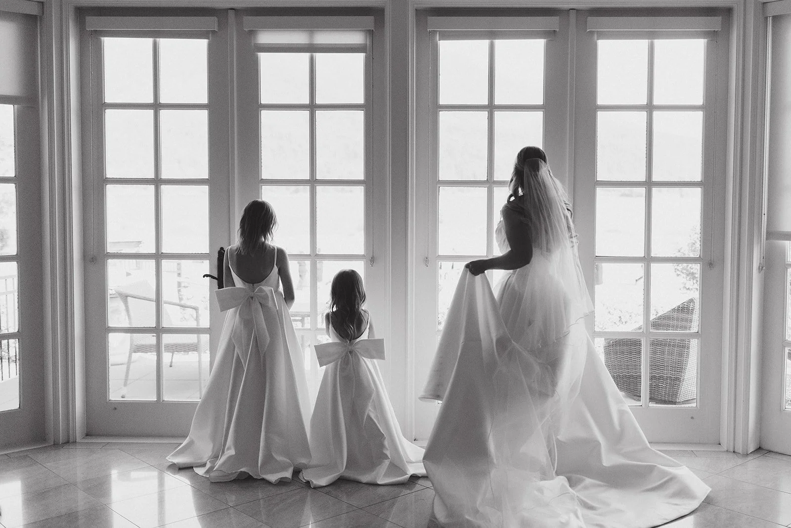 Three women and a girl in white dresses with large bows, standing by large French doors, looking outside. One woman holds up her dress, and the girl and the other woman stand with their backs to the camera.