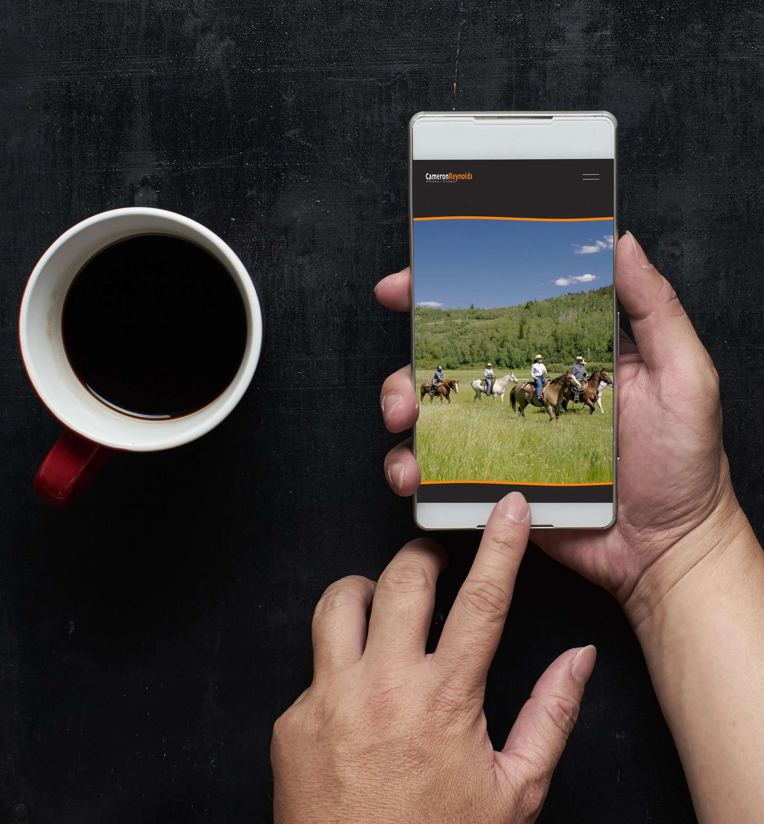 Hands holding a cell phone looking at Cameron Reynolds Photography om the phone screen with a cup of coffee next to them.