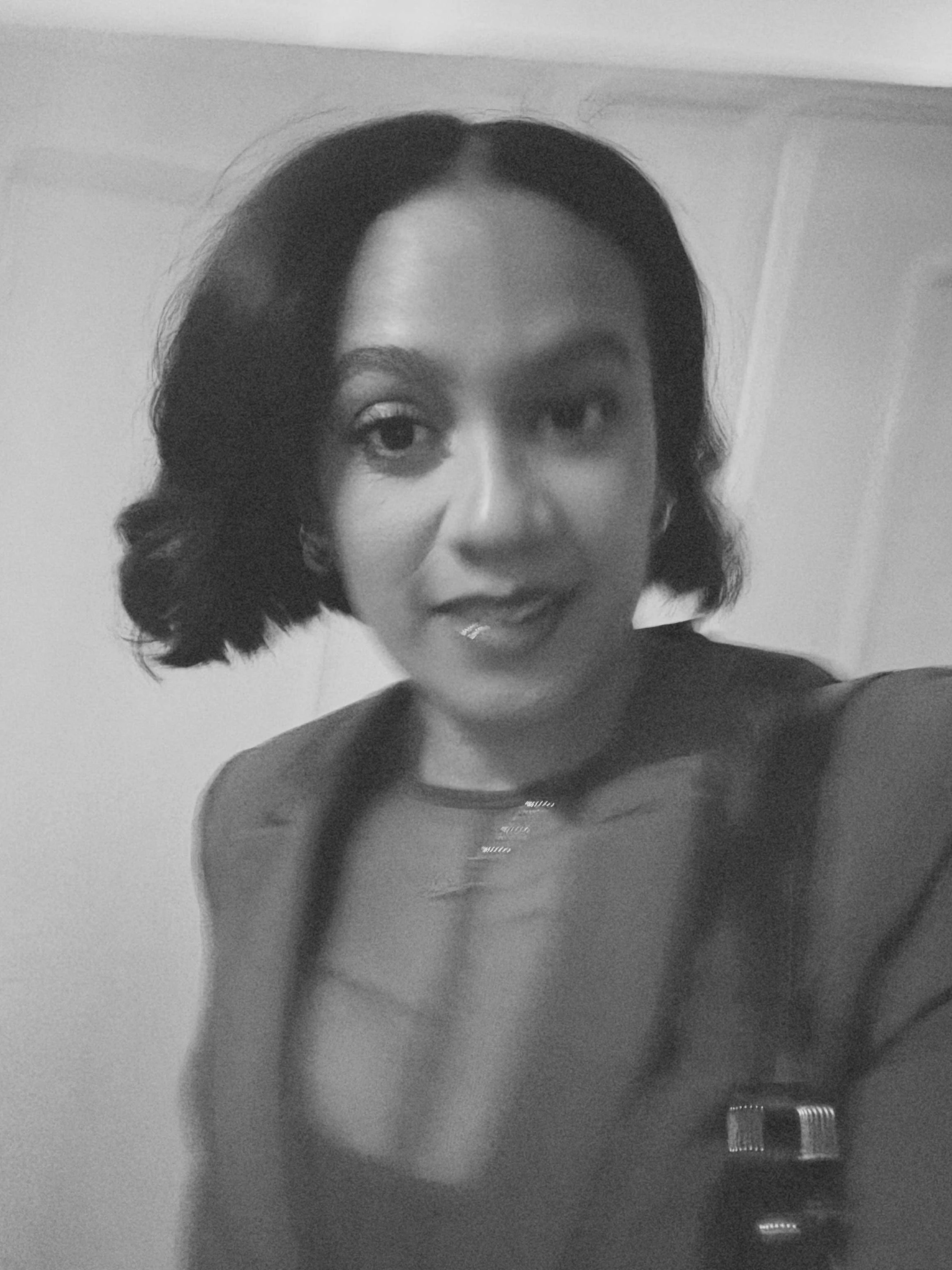 A woman with short, dark, wavy hair and medium skin tone takes a selfie in black and white, with her face partially visible in the frame. She is standing in front of a plain wall and a mirror, and her shoulder is visible in the lower part of the image.