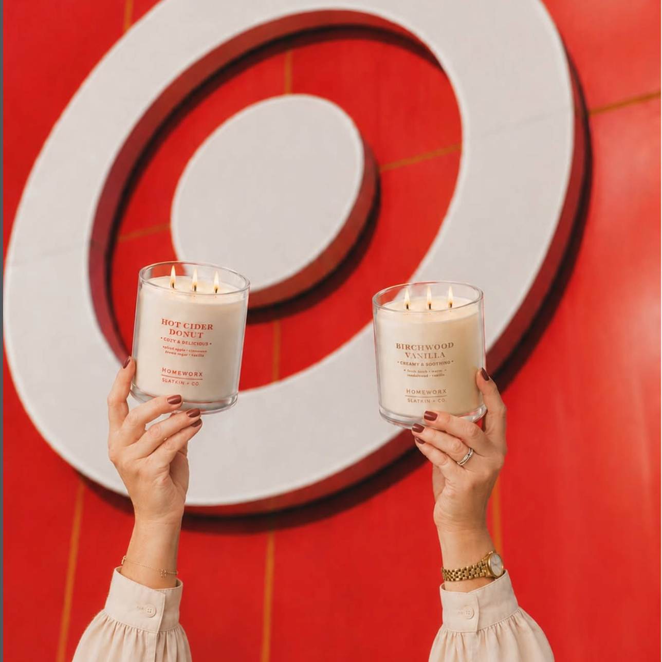 Two hands holding white candles with flaming wicks in front of a red and white target wall.