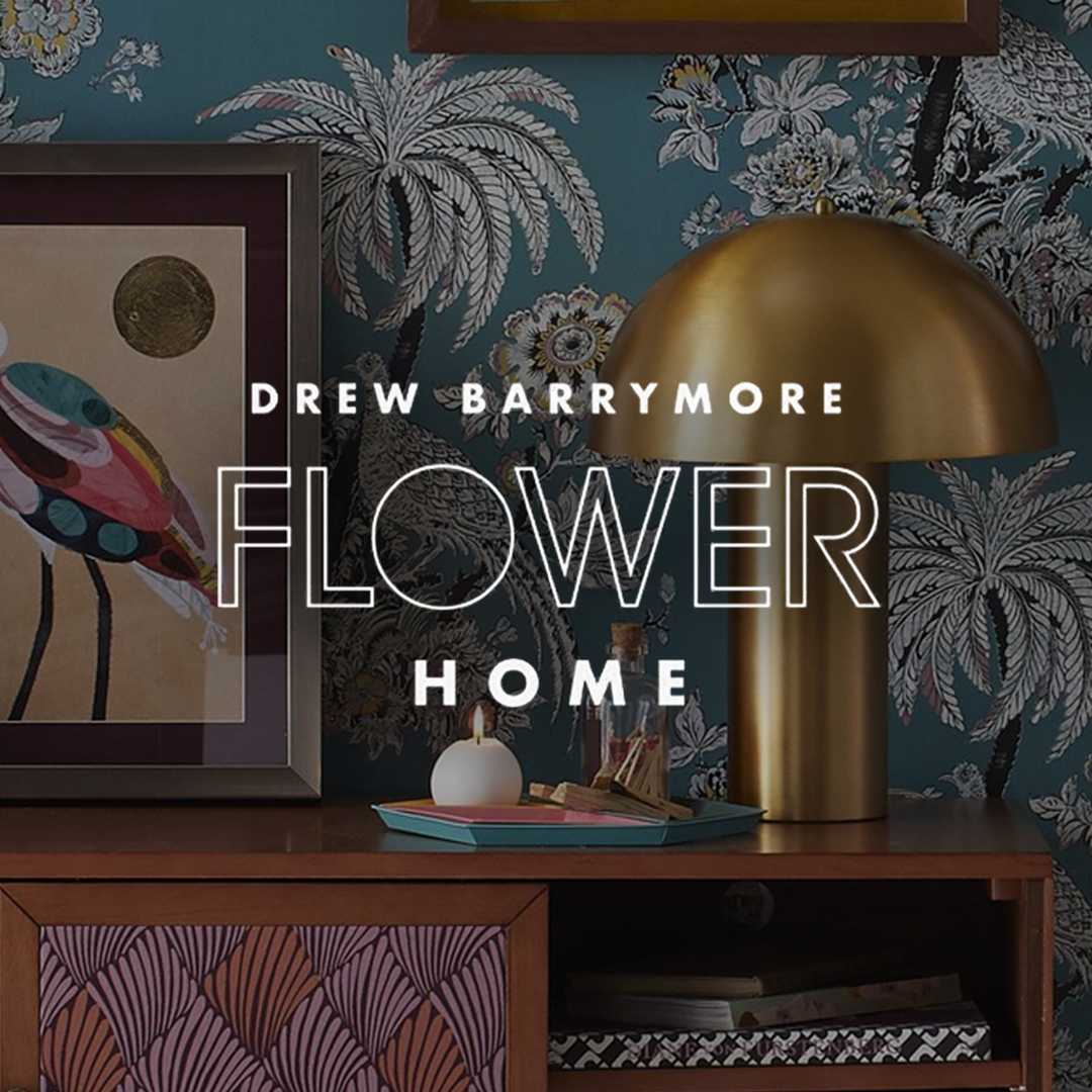 A home decor scene with a framed artwork of a colorful insect, a gold dome-shaped table lamp, a small lit candle, and decorative objects on a wooden surface. The background features teal wallpaper with white and black floral and palm tree patterns.