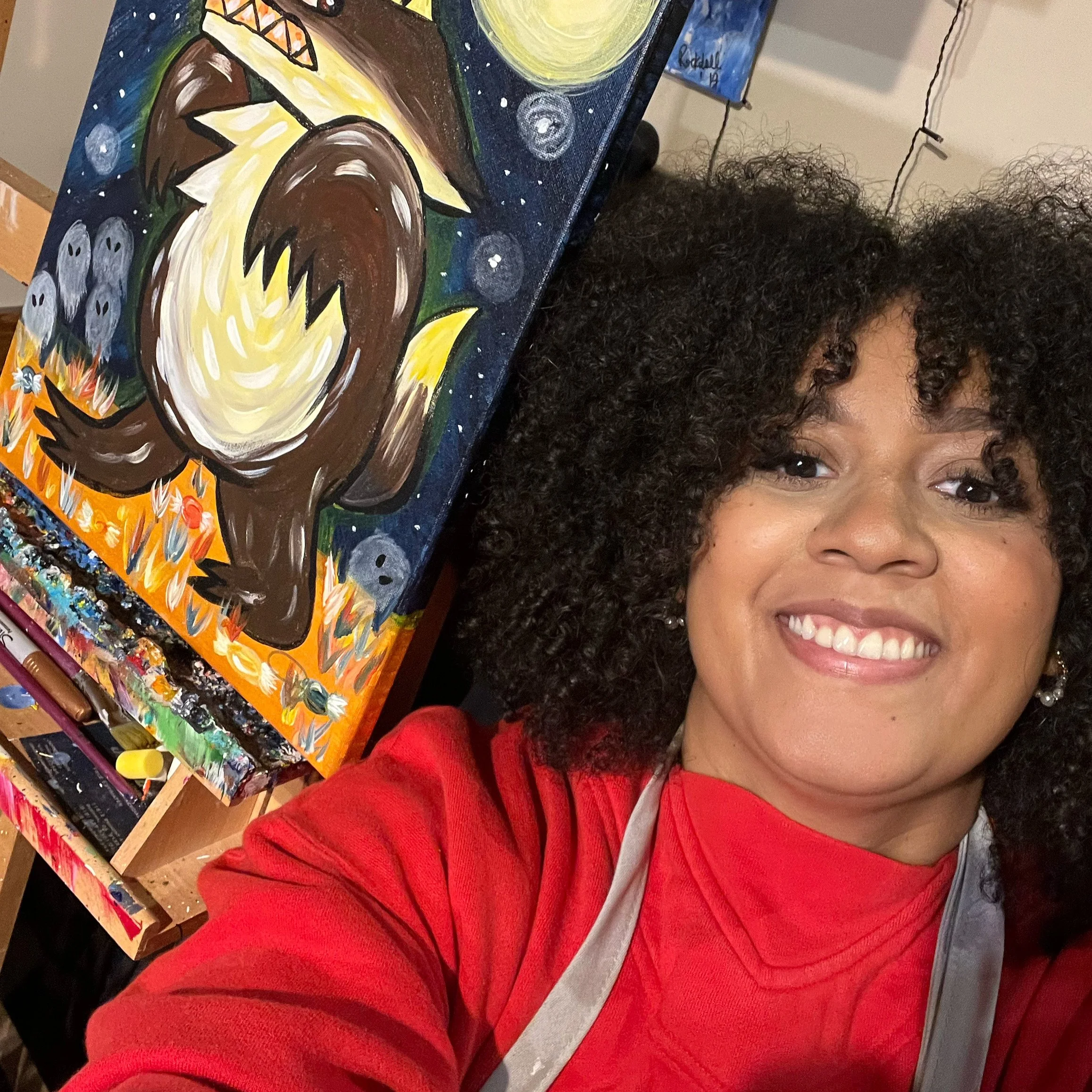 Close-up of a woman with dark, curly hair smiling, wearing a red sweater and white shirt, with an abstract painted canvas featuring a large yellow moon, stars, and a colorful landscape in the background.