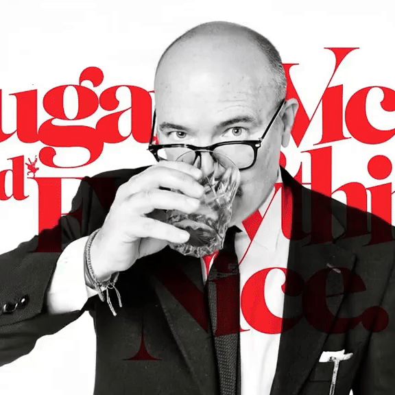 A man with glasses in a suit drinks from a glass of dark beverages, with artwork and text in red behind him.