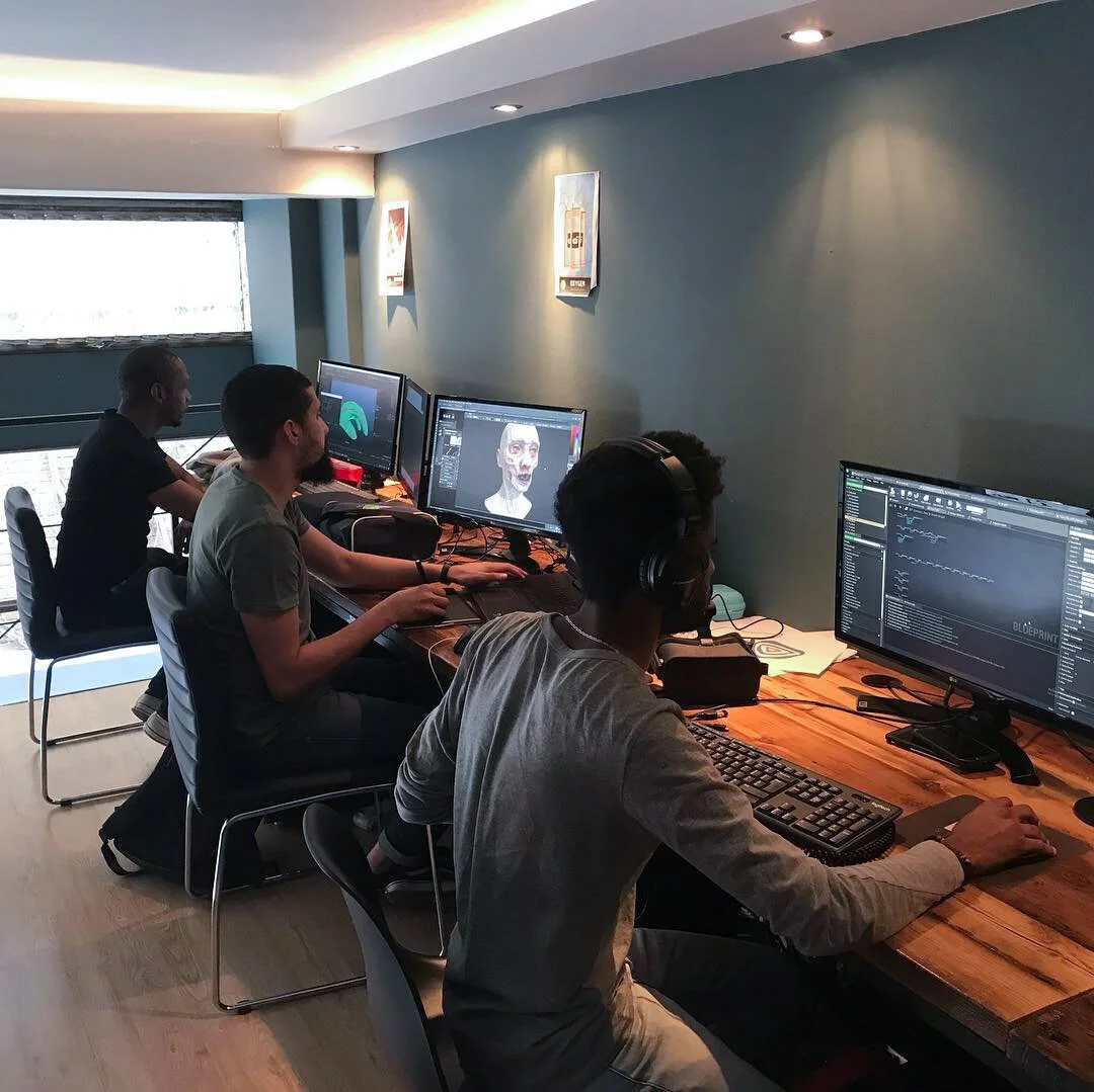 Four people working on computers at a long wooden desk in a room with teal walls. The monitors display digital art and sound editing software, with one showing a 3D model of a human face. Sunlight streams in through a window, and framed artwork hangs on the wall.