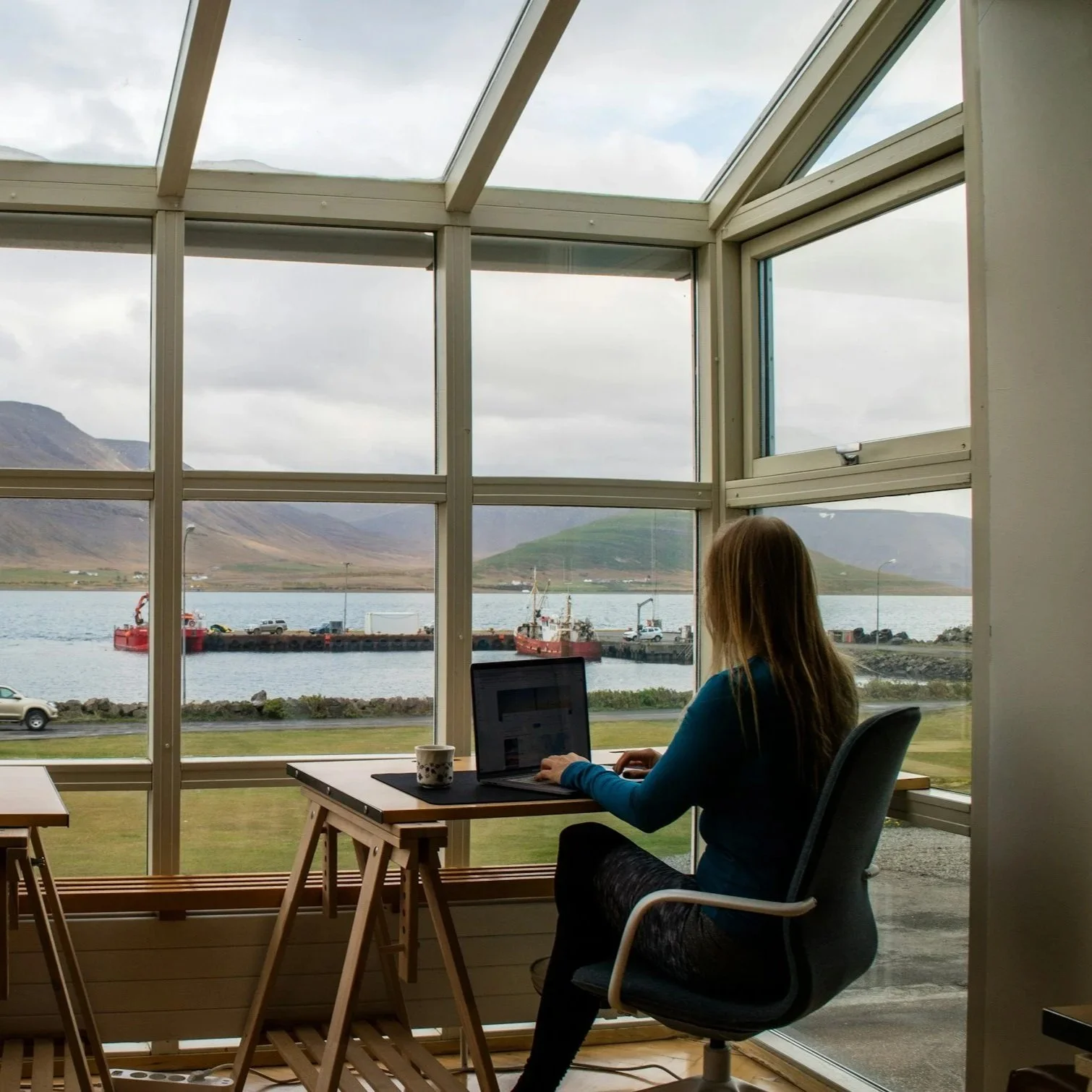 A woman sitting at a desk working on a laptop in a room with large windows overlooking a harbor with ships, mountains, and a cloudy sky.