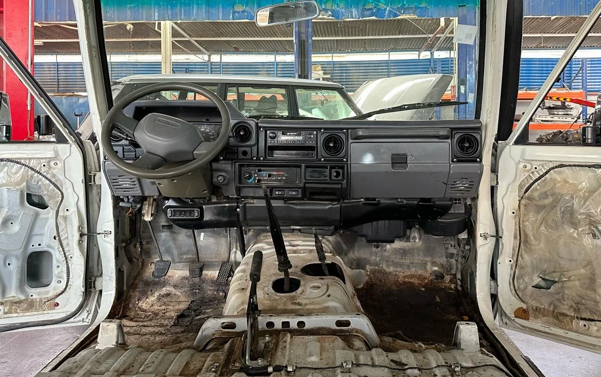 Stripped interior of a Toyota Land Cruiser 78 Hardtop in a workshop with seats, carpet, and door panels removed, showing the bare floor pan, pedals, gear levers, and dashboard