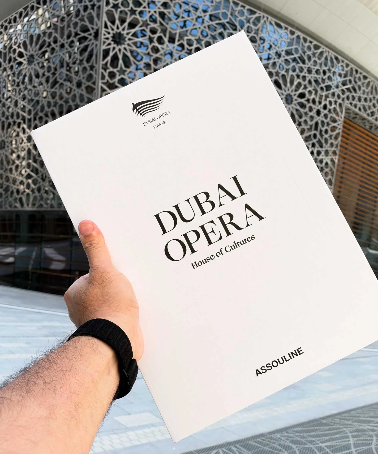 The Assouline book Dubai Opera: House of Cultures, featuring architectural photography by Dany Eid, held in front of Dubai Opera's geometric façade in Downtown Dubai.