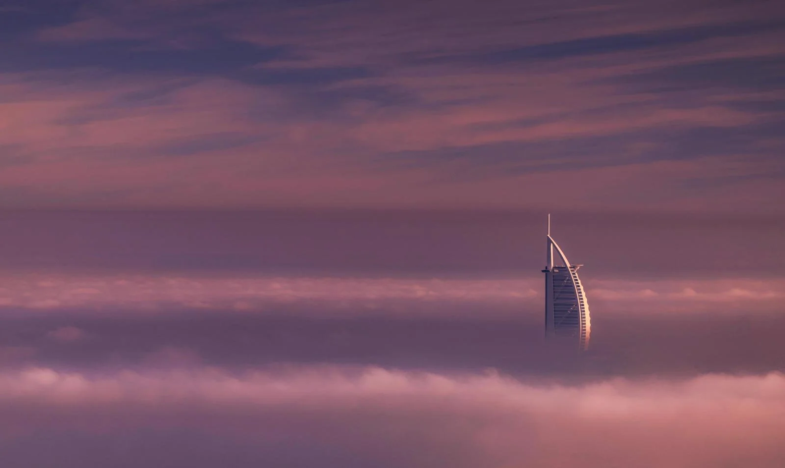 Burj Al Arab tower rising above a layer of fog and clouds under a pink and purple sky in Dubai