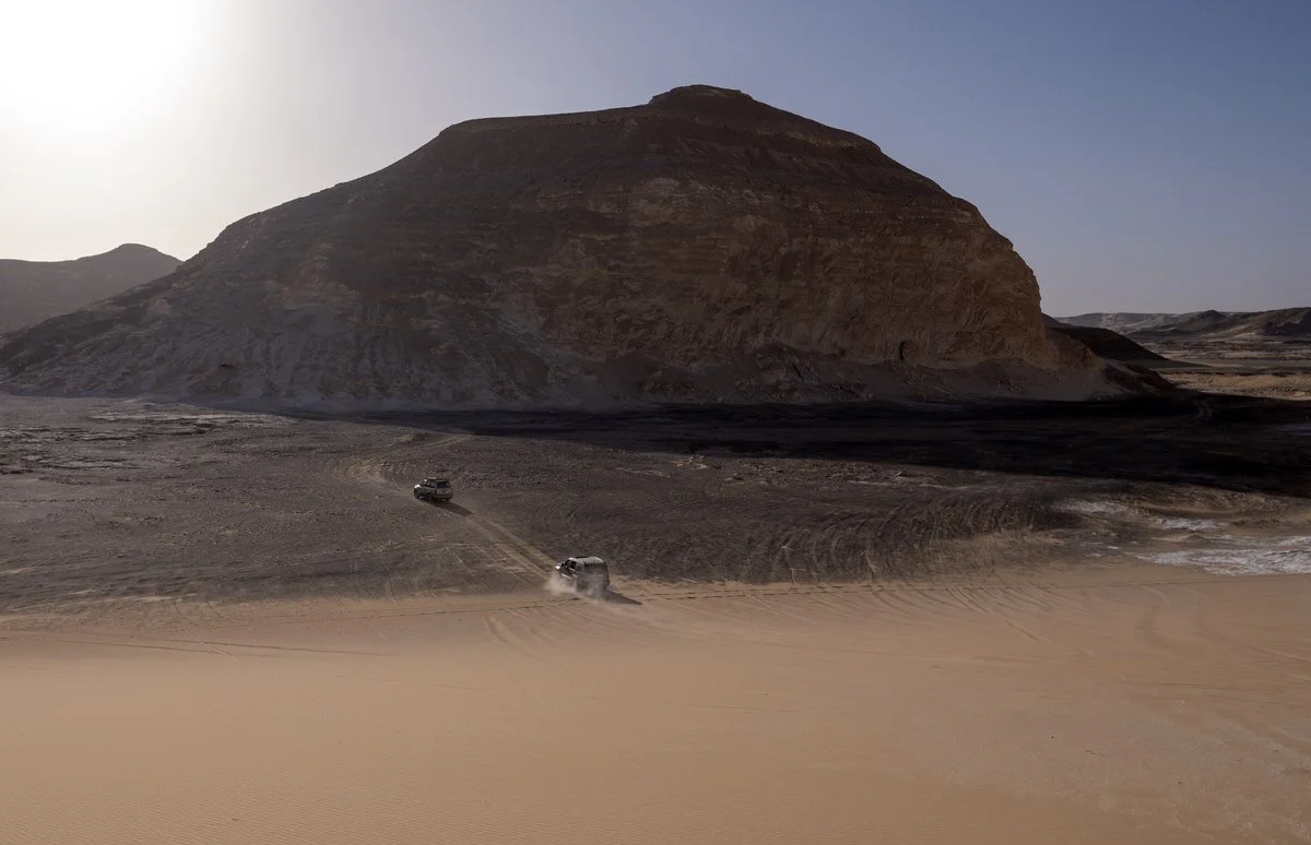Two 4x4 vehicles crossing sandy terrain beneath a towering rock formation in Egypt’s Western Desert