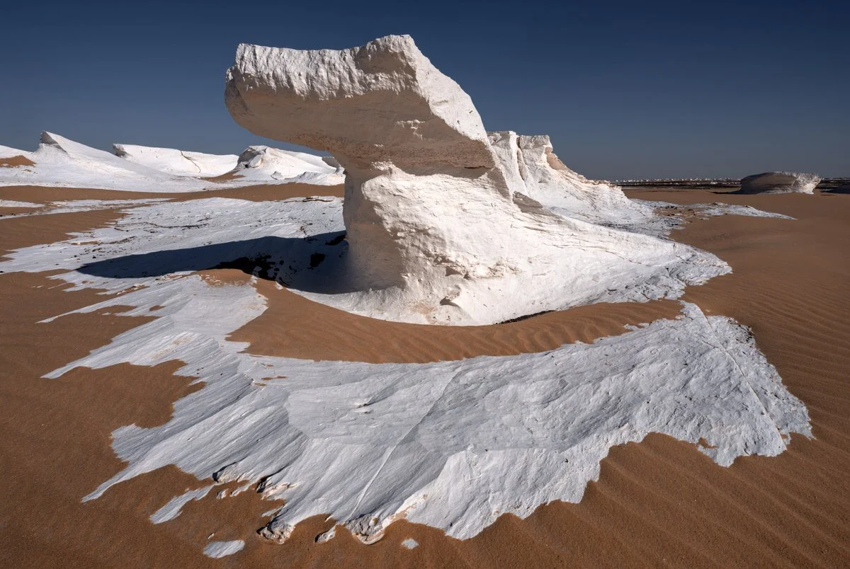 A wind-sculpted chalk formation rises above rippled golden sand beneath a deep blue sky in Egypt’s White Desert, photographed by Dany Eid.