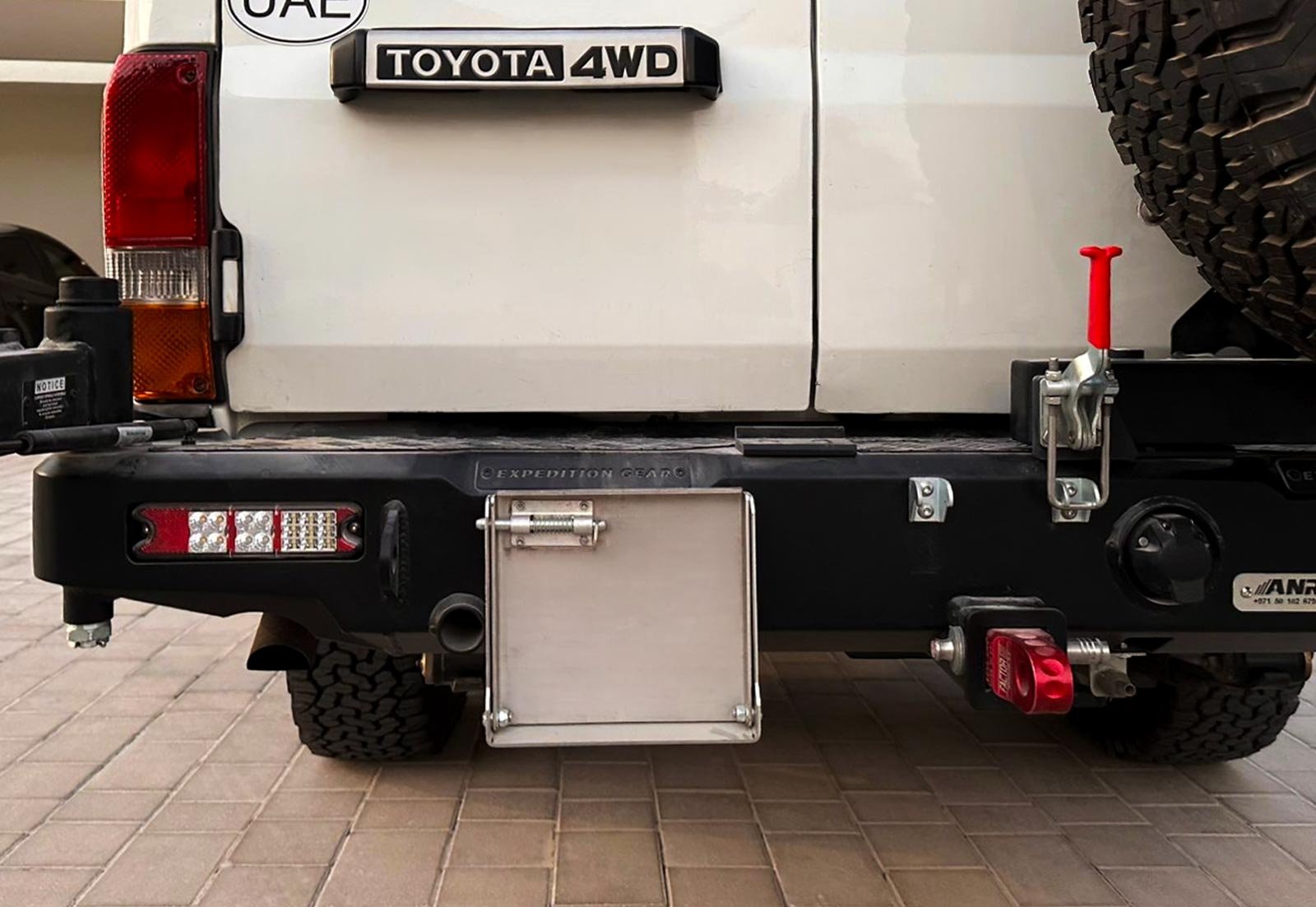 Rear of a white Toyota Land Cruiser 78 Hardtop with a black heavy-duty rear bumper, LED lights, red recovery shackle, spare tire carrier latch, and aluminium license plate holder