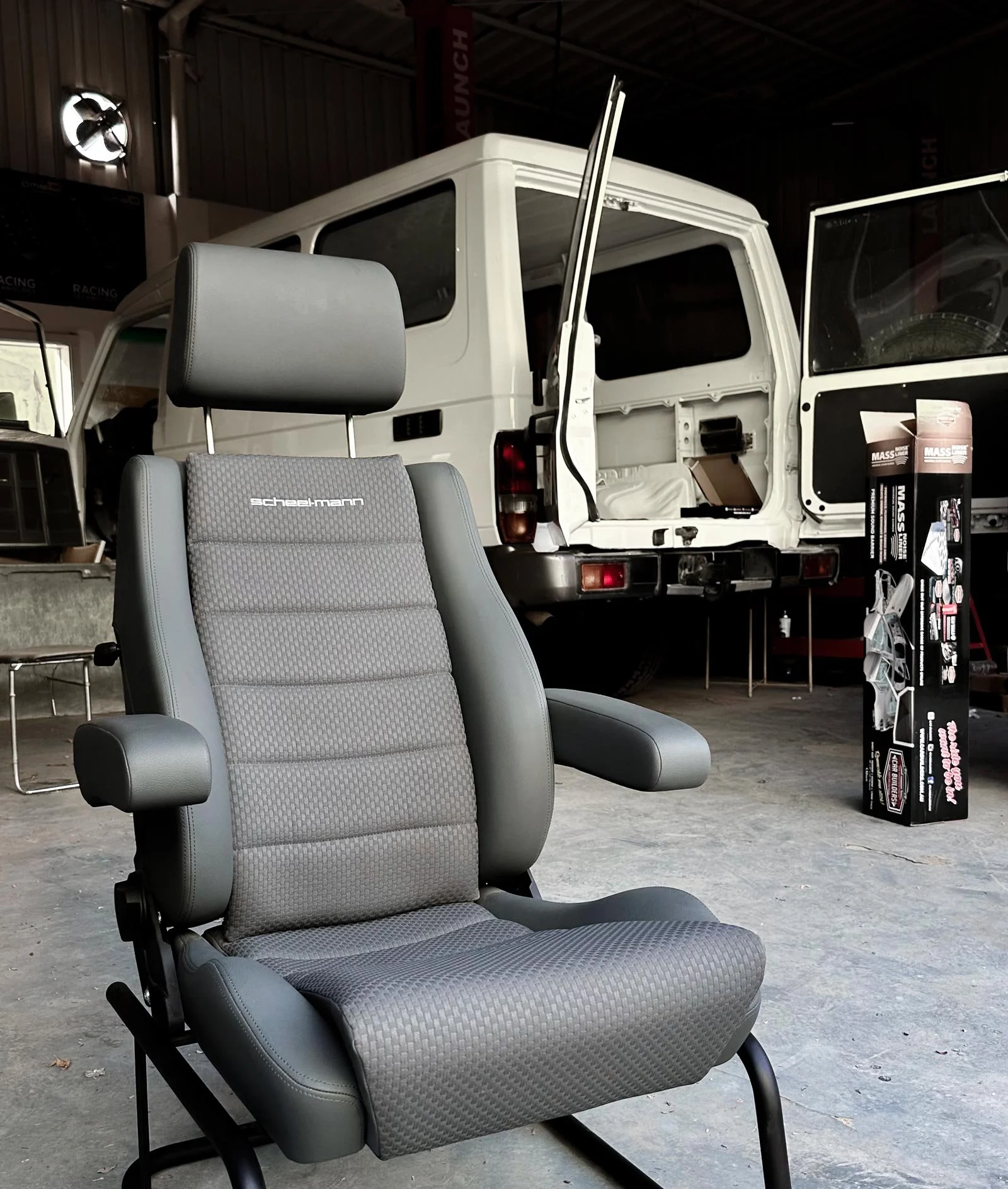New grey Scheel-Mann seat with armrests in a workshop, with a repainted white Toyota Land Cruiser 78 Hardtop behind it and a box of Mass Noise Liner sound insulation beside it