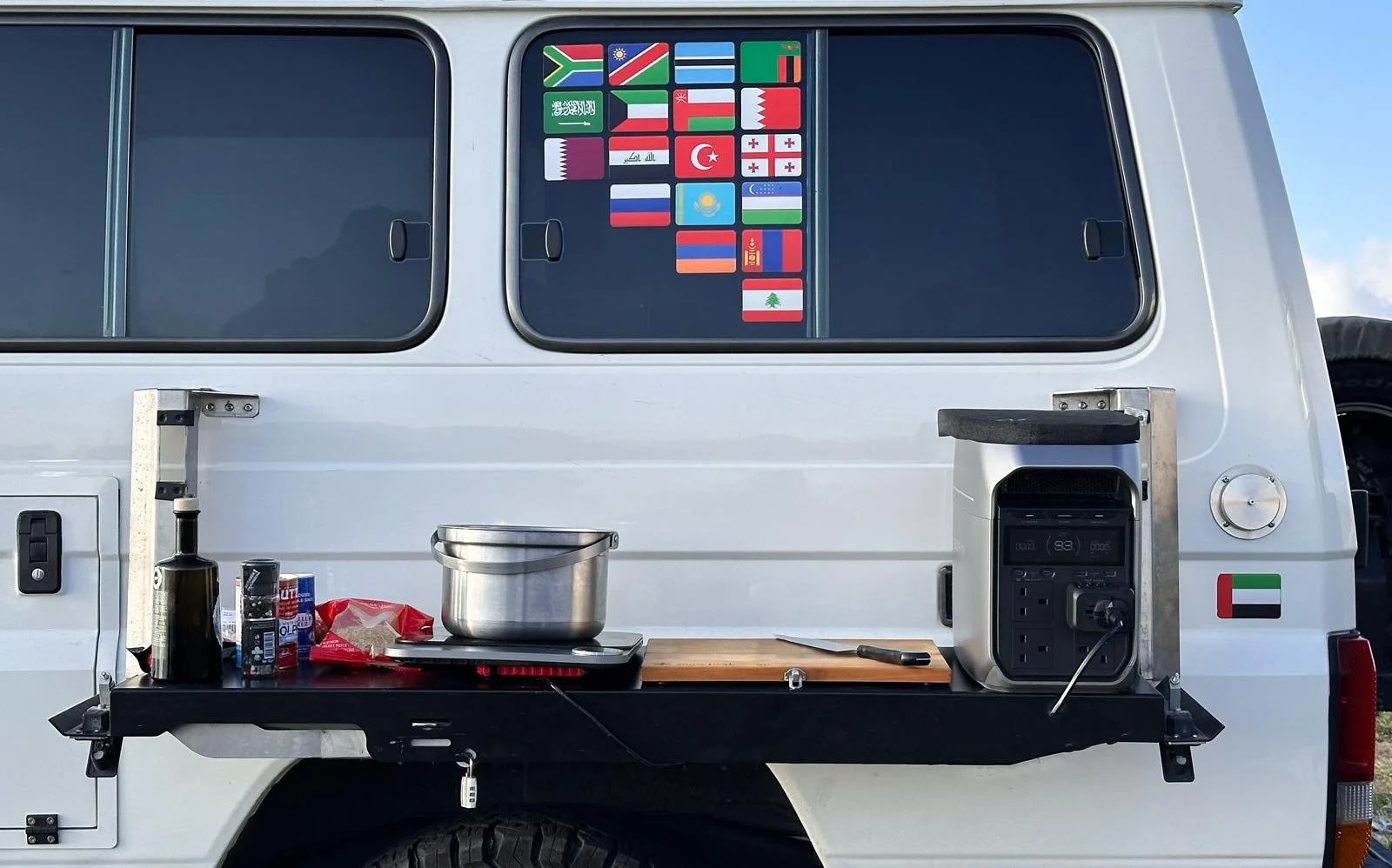 White Toyota Land Cruiser 78 Hardtop with a custom side mount that holds MaxTrax recovery boards and doubles as a side table with a pot, induction cooktop, cutting board, and portable power station