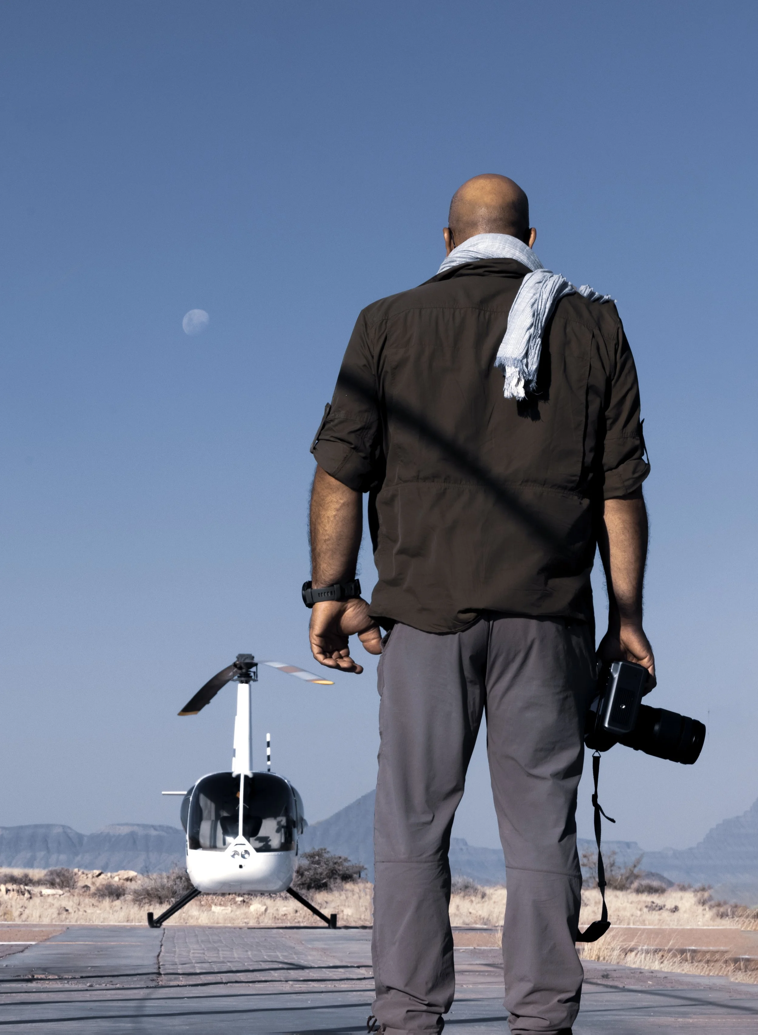 Photographer holding a camera on location for an aerial photography assignment, facing a helicopter on a desert helipad in Namibia