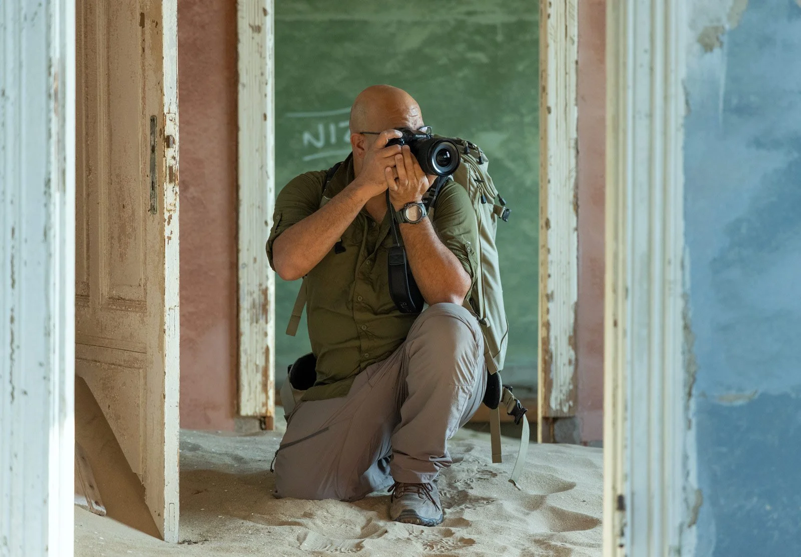 Photographer kneeling in sand with a Fujifilm camera and backpack between weathered doorframes in Kolmanskop, Namibia.