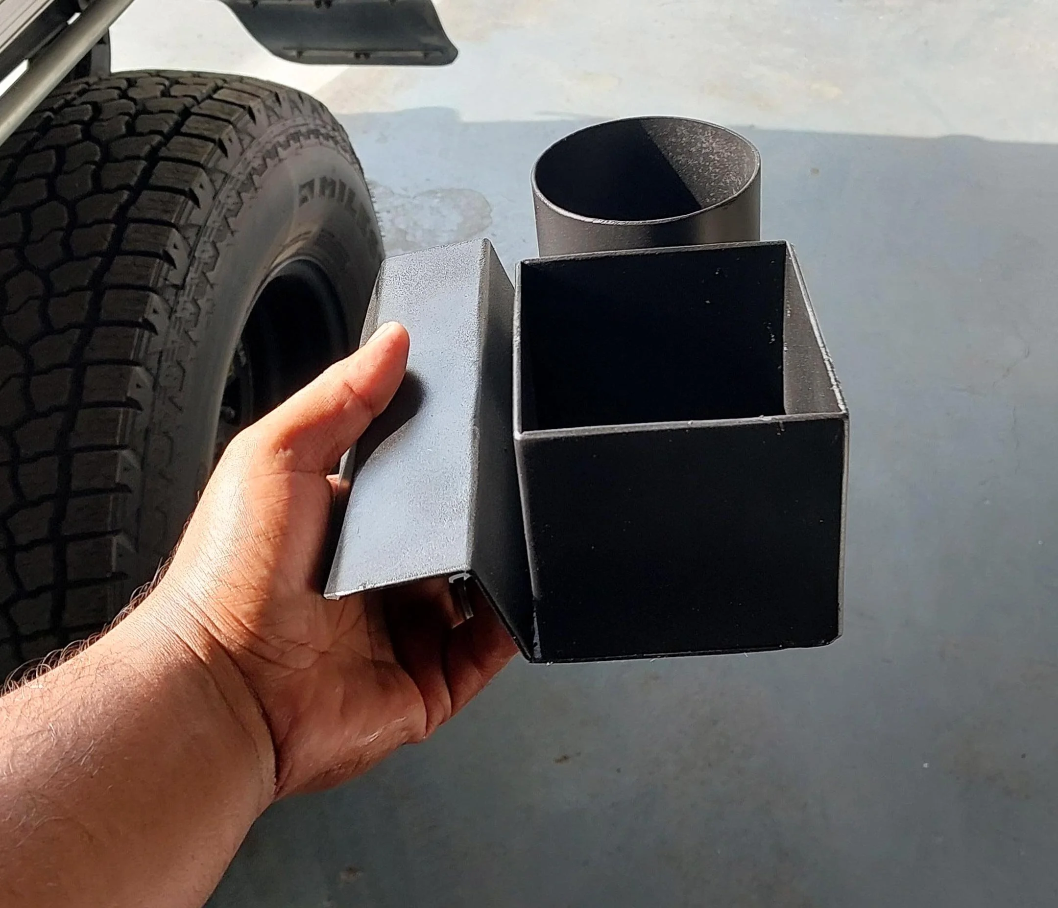Hand holding a custom black powder-coated steel cup holder with a hinged lid and round tube insert next to the tire of a Toyota Land Cruiser 78