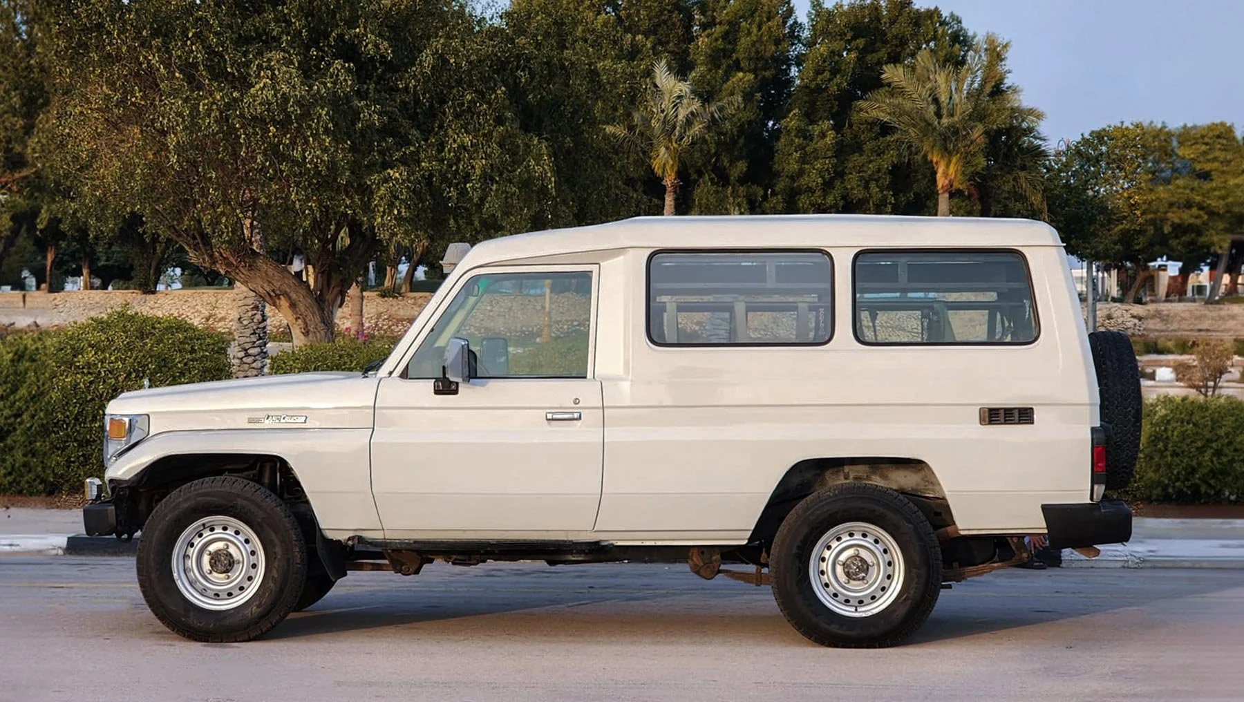 Side view of a white Toyota Land Cruiser 78 Hardtop with steel wheels and a rear spare tire, parked on a road with palm trees and greenery behind it