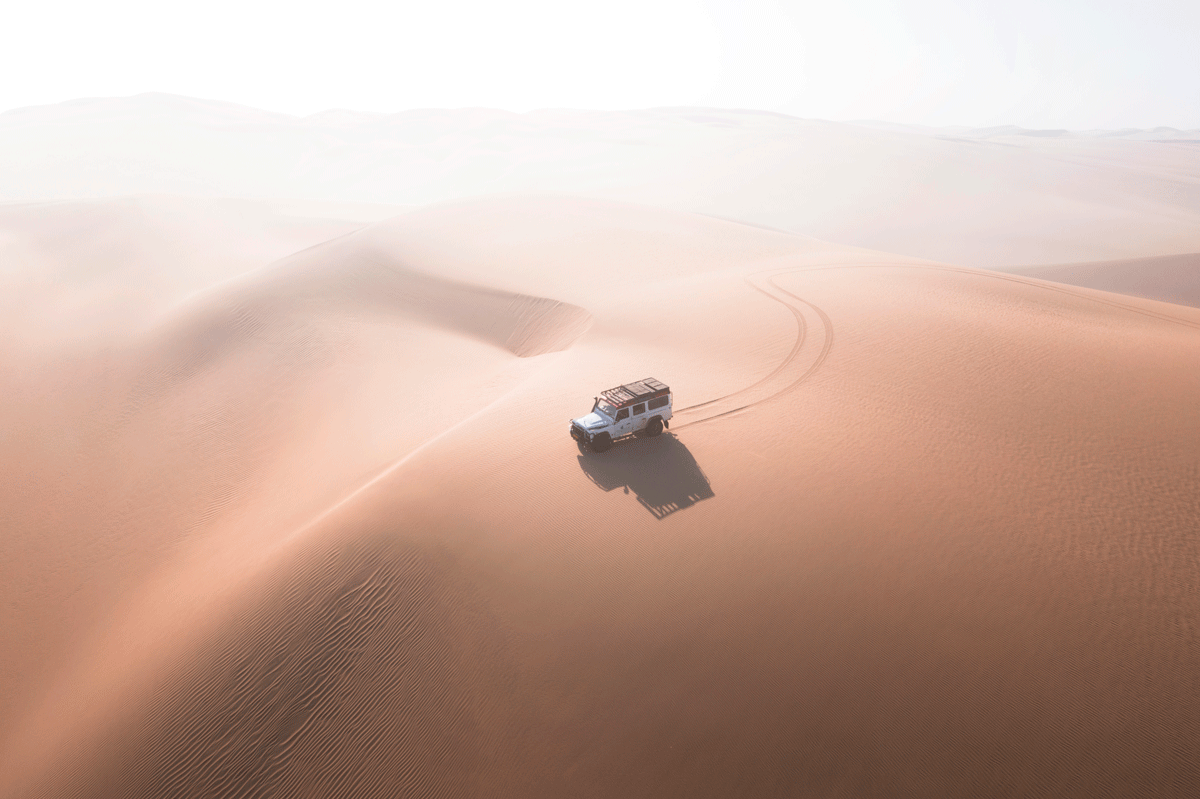 Aerial view of a 4x4 expedition vehicle leaving tracks across a soft pink sand dune in Namibia