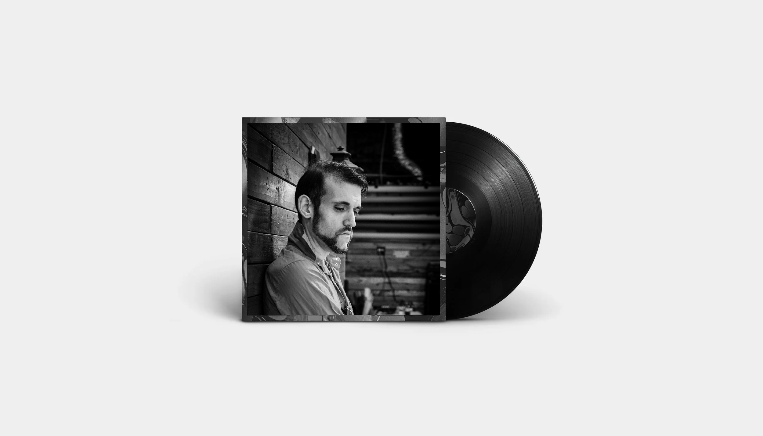 Black and white album cover featuring a man with a beard and mustache looking down, sitting against a wooden wall in a studio setting, with a vinyl record partially visible behind the cover.