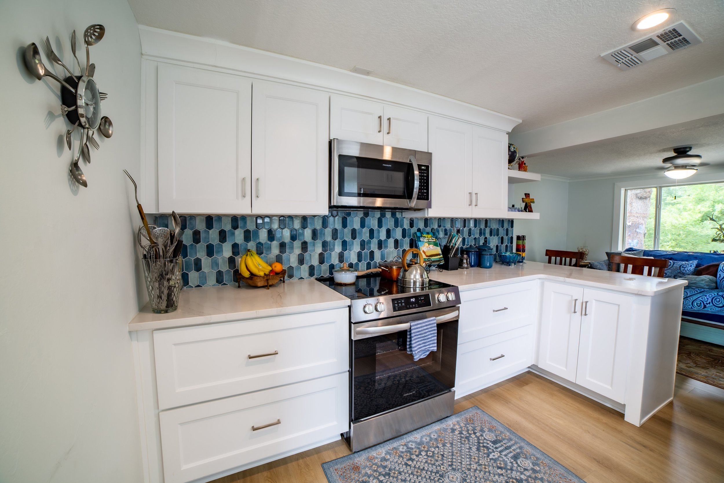 A bright kitchen featuring white shaker cabinets, light quartz countertops, stainless steel appliances, and a bold blue hexagonal tile backsplash, opening to a sunlit living and dining area.