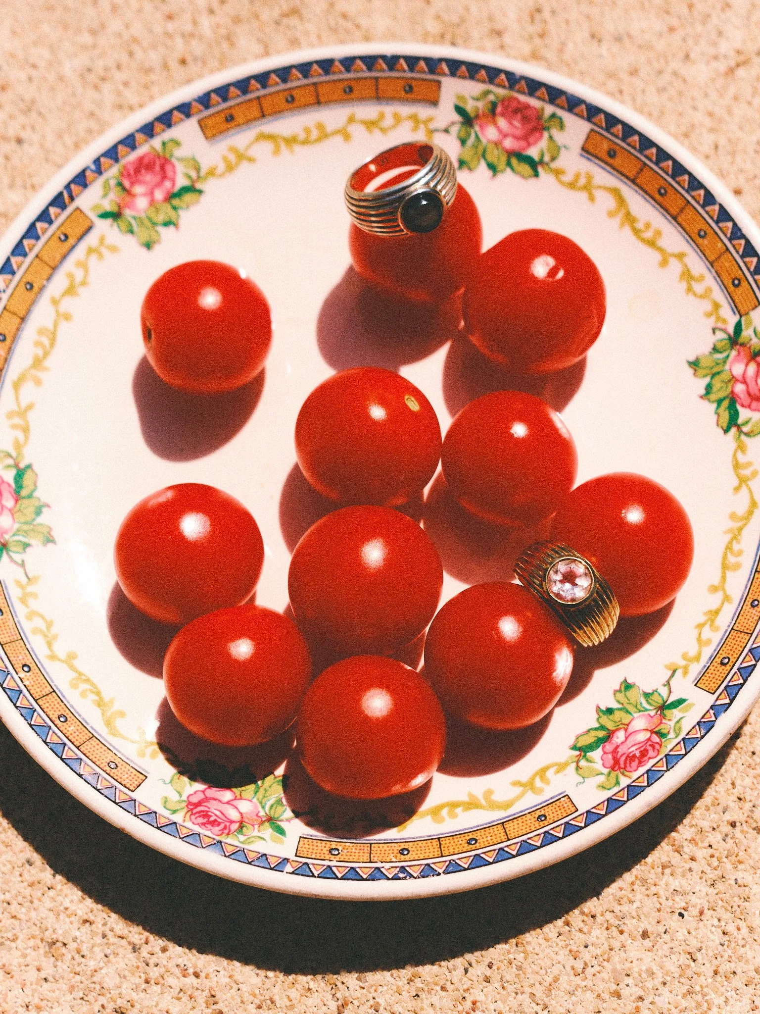 `Editorial jewelry still life with vintage rings and red tomatoes on a floral plate, photographed in bright Mediterranean summer sunlight