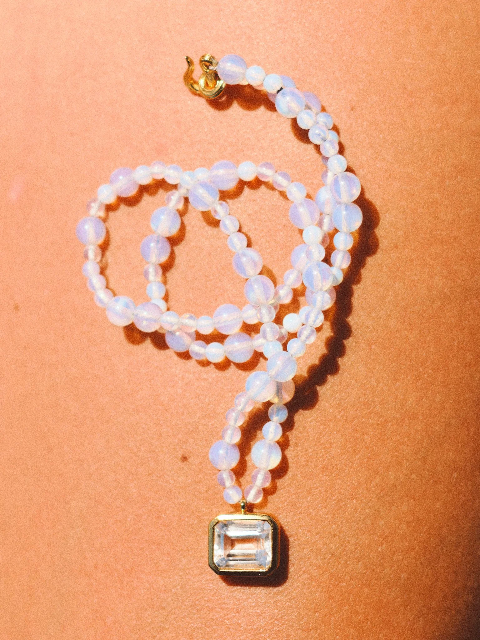 `Editorial jewelry still life of an opalescent beaded necklace with crystal pendant on sun-kissed skin, photographed in warm Mediterranean light
