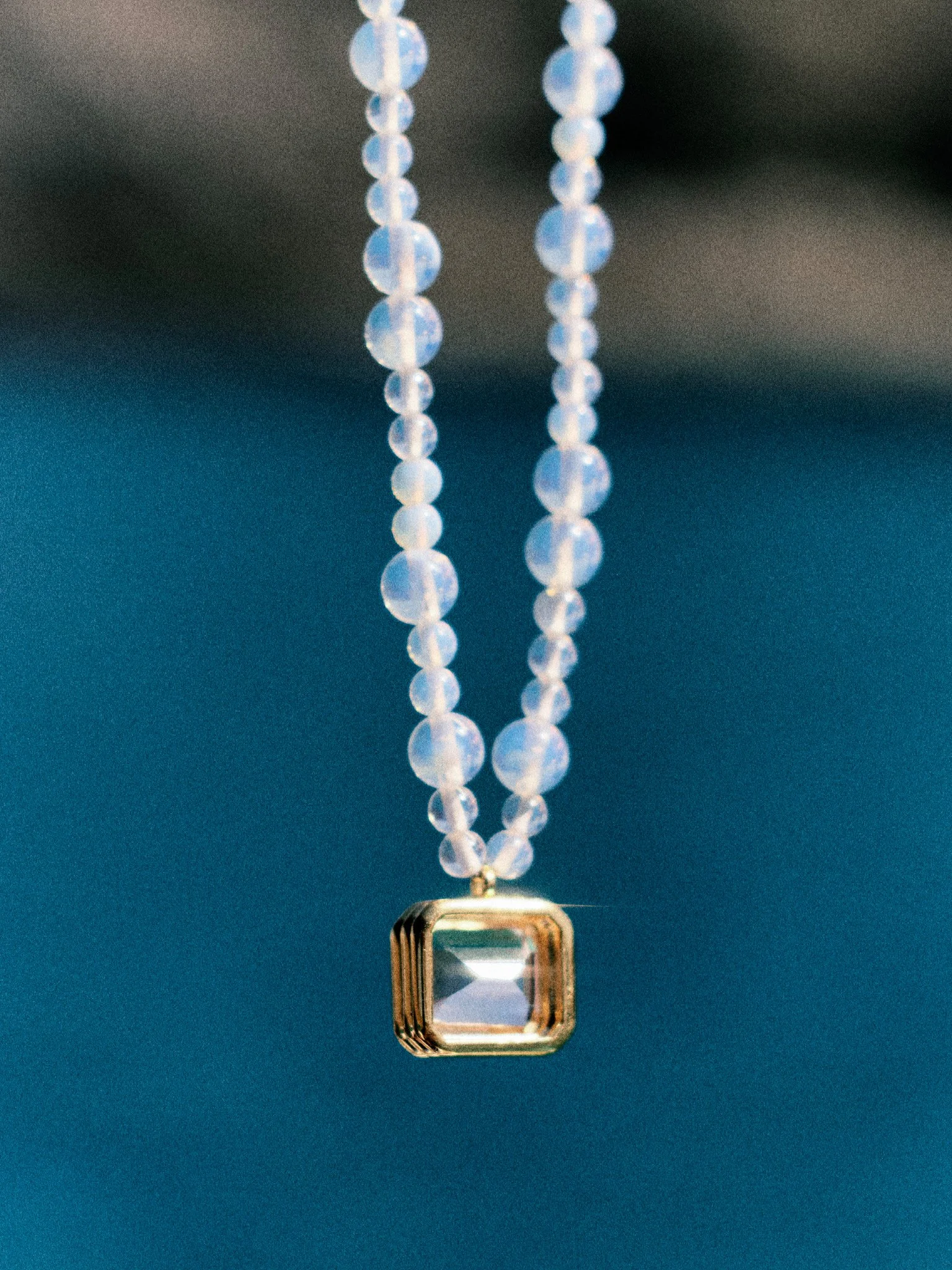 `Editorial jewelry still life of an opalescent beaded necklace with crystal pendant against a deep blue Mediterranean background.