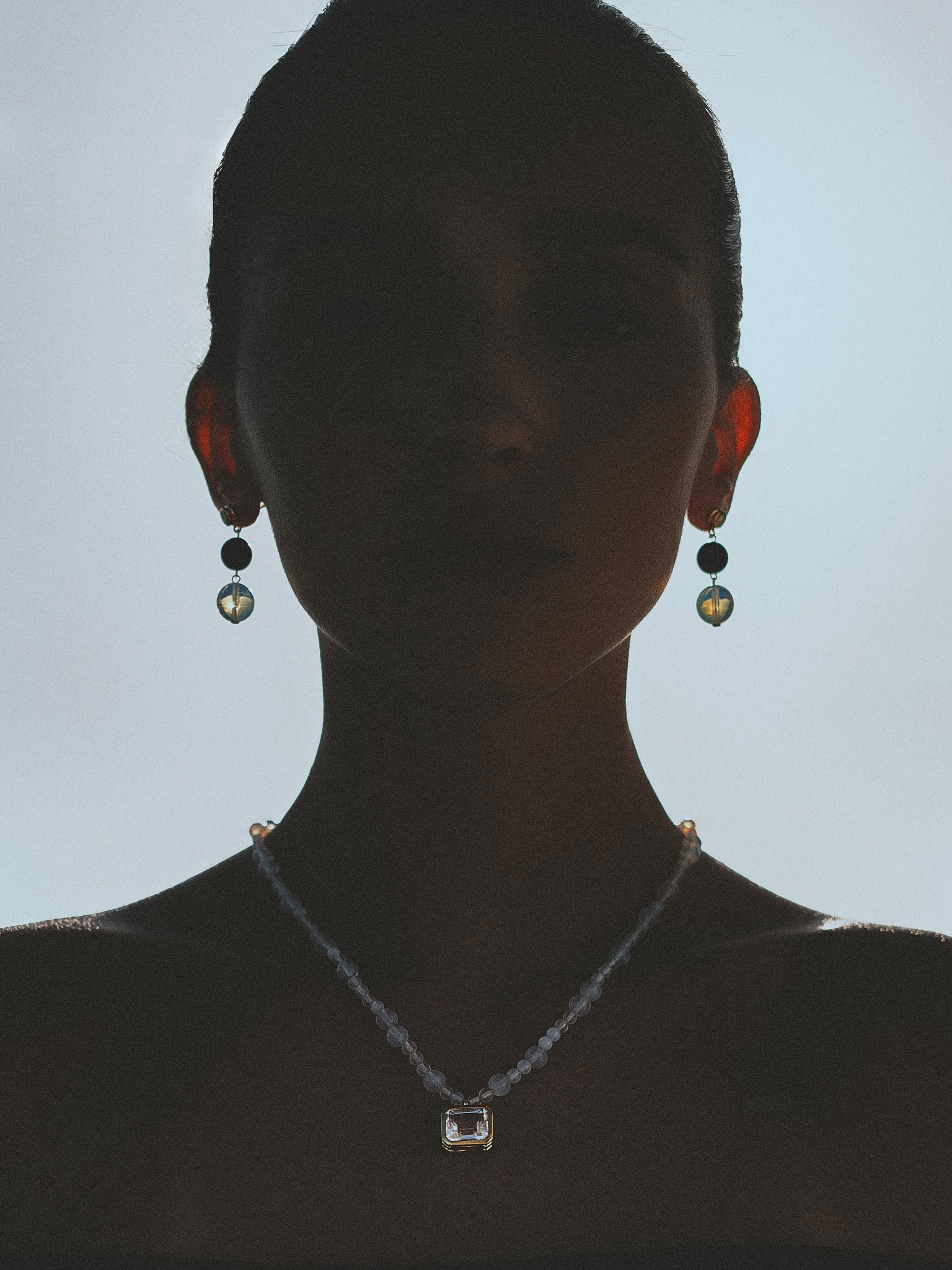 `Editorial jewelry portrait in silhouette wearing crystal earrings and a beaded pendant necklace, photographed in soft backlight.