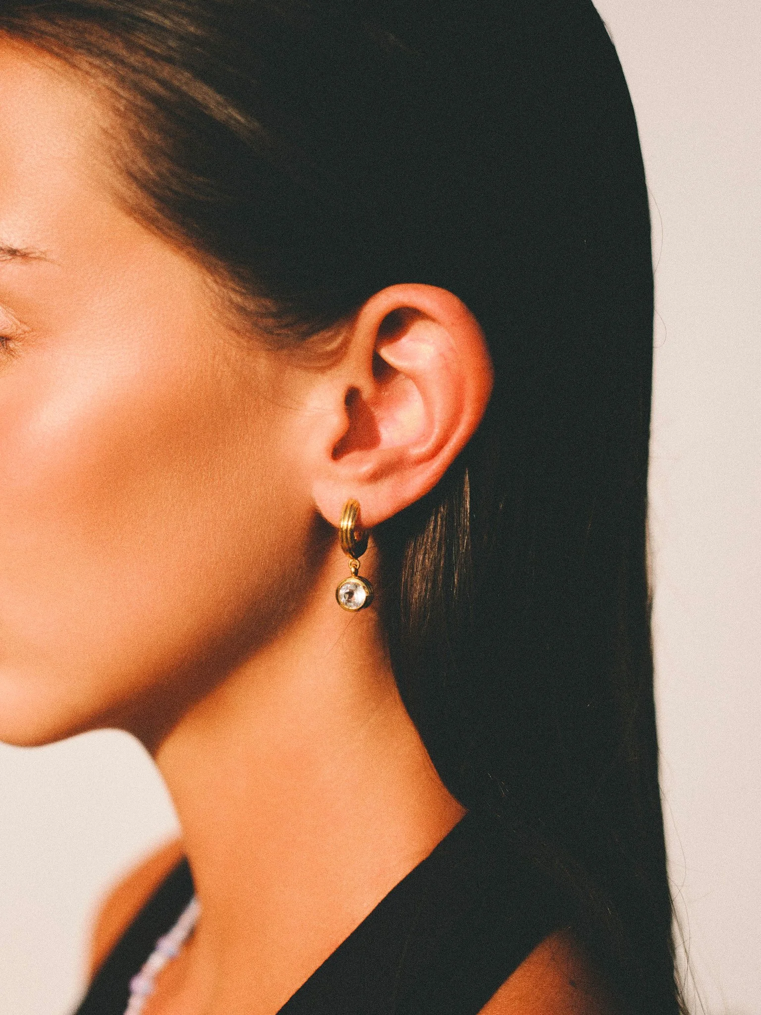 `Editorial jewelry close-up of a woman wearing a gold hoop earring with crystal pendant, photographed in warm natural light.