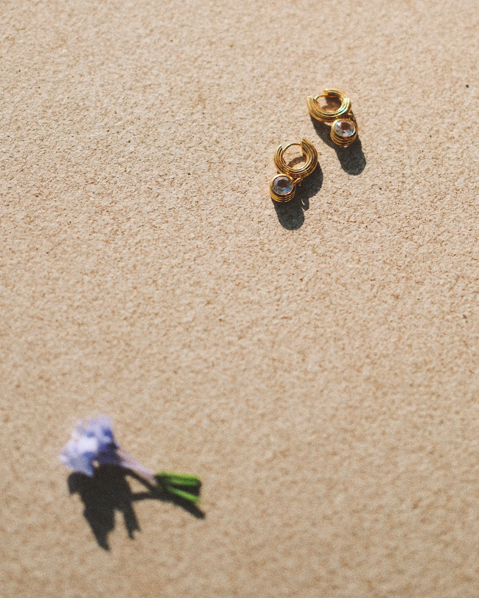 `Editorial jewelry still life of gold and crystal earrings on a sunlit stone surface with a small purple flower, photographed in Mediterranean summer light.