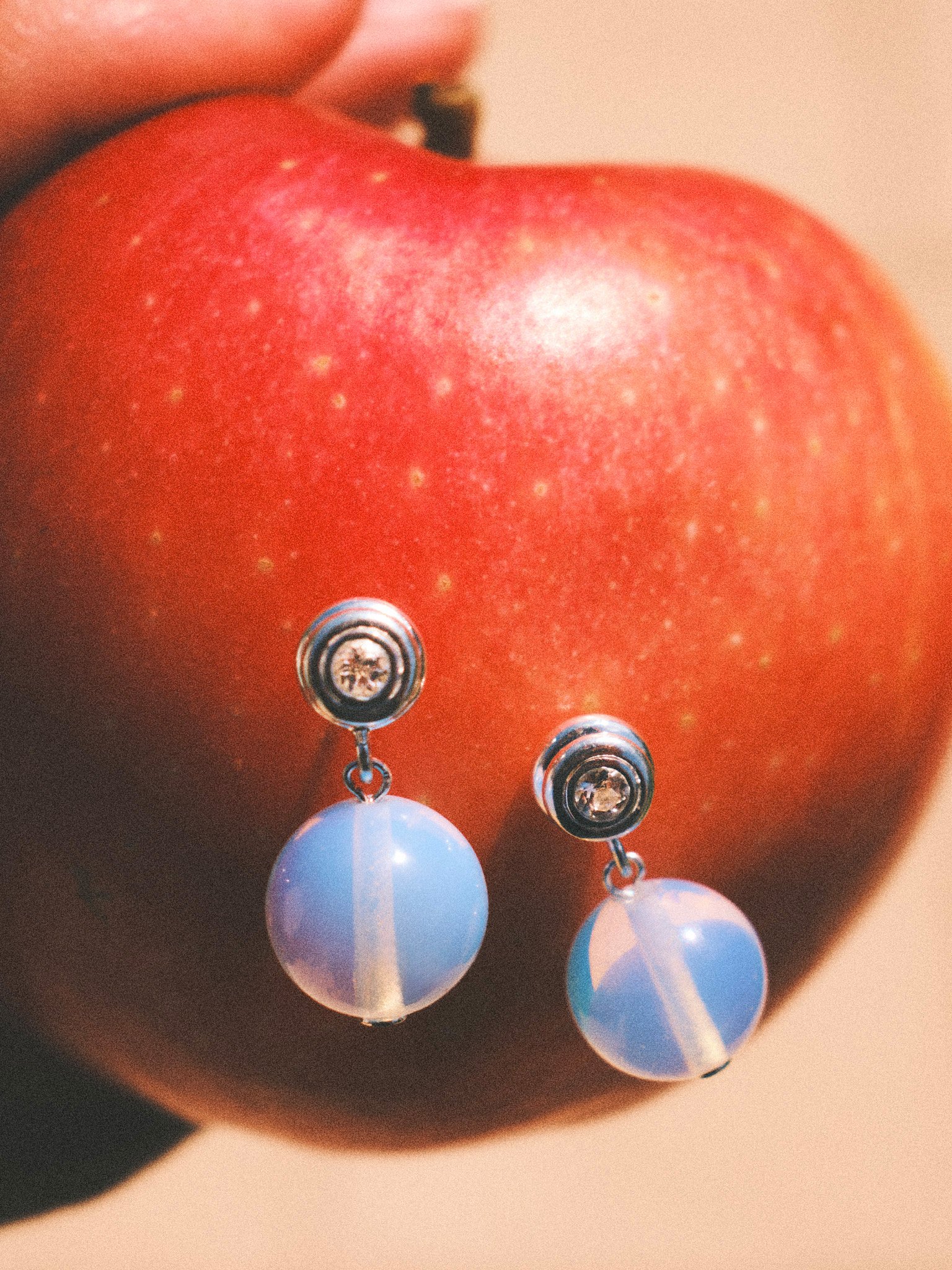 `Editorial jewelry still life of opalescent drop earrings styled on a red apple, photographed in warm natural light.