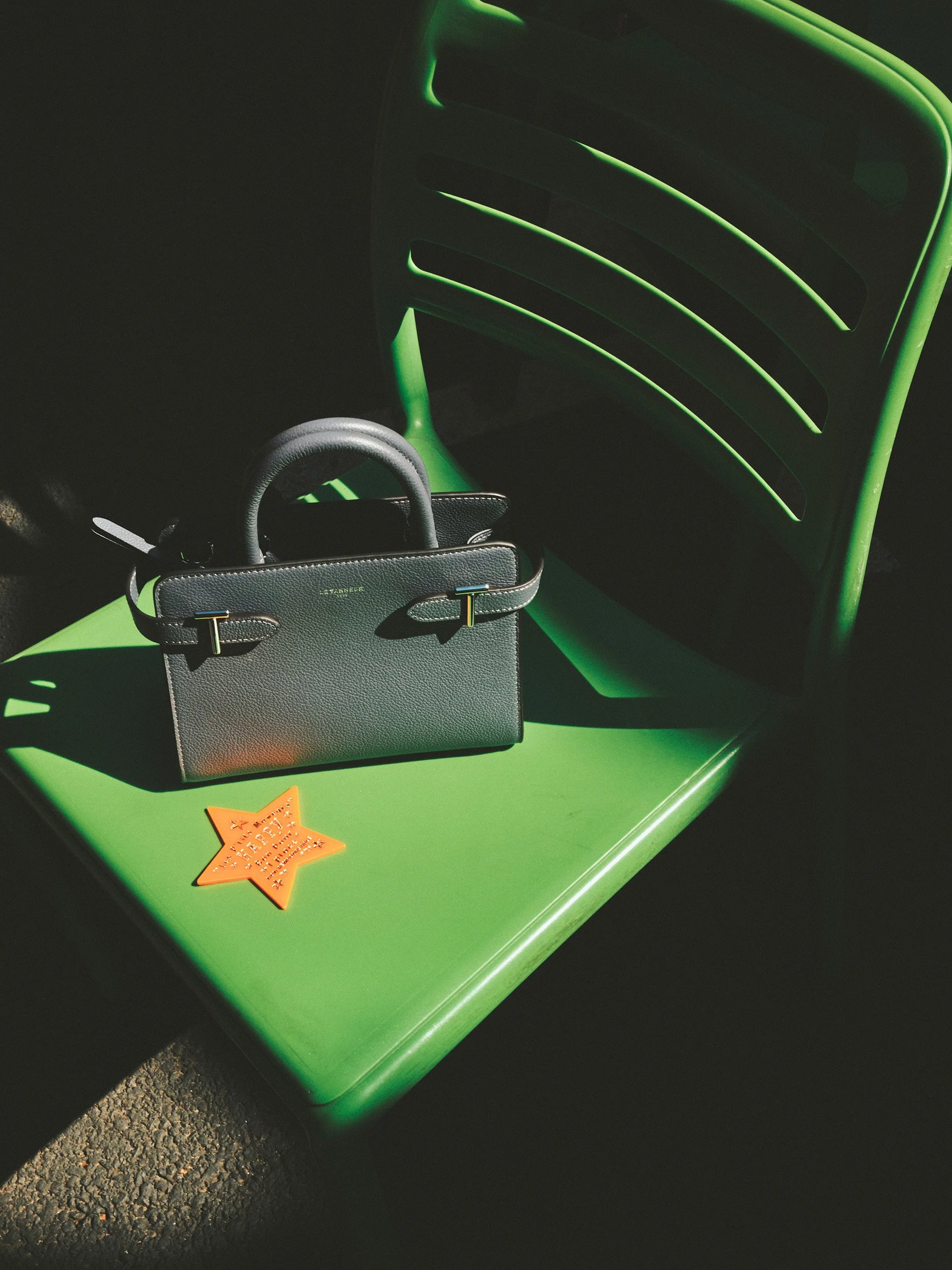 Editorial fashion still life of a black Le Tanneur leather handbag on a bright green chair, photographed in dramatic sunlight and shadow.