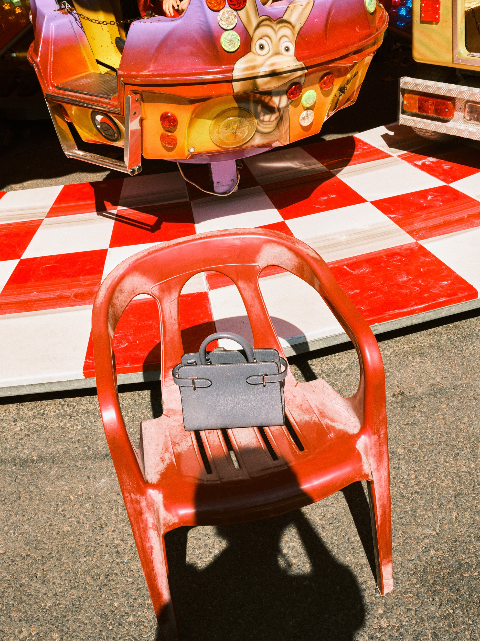 Editorial fashion still life of a small Le Tanneur leather handbag on a red plastic chair at a colorful fairground, photographed in bright summer sunlight.